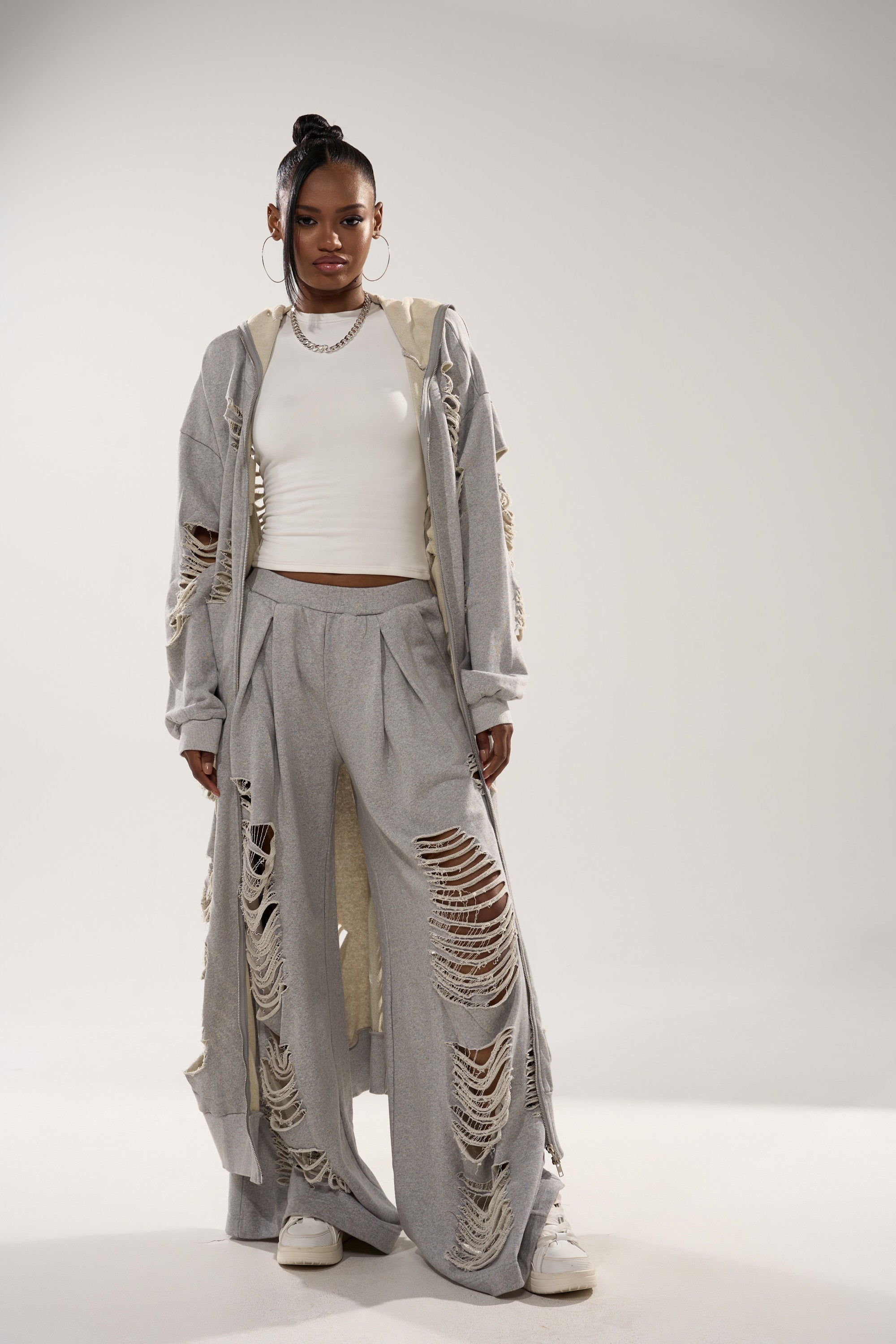 A woman exudes confidence in the STRESS HIM OUT LONGLINE SWEATSHIRT, a light gray, distressed long coat paired with matching wide-leg pants, a white crop top, sneakers, hoop earrings, and a silver chain necklace against a plain backdrop. 2