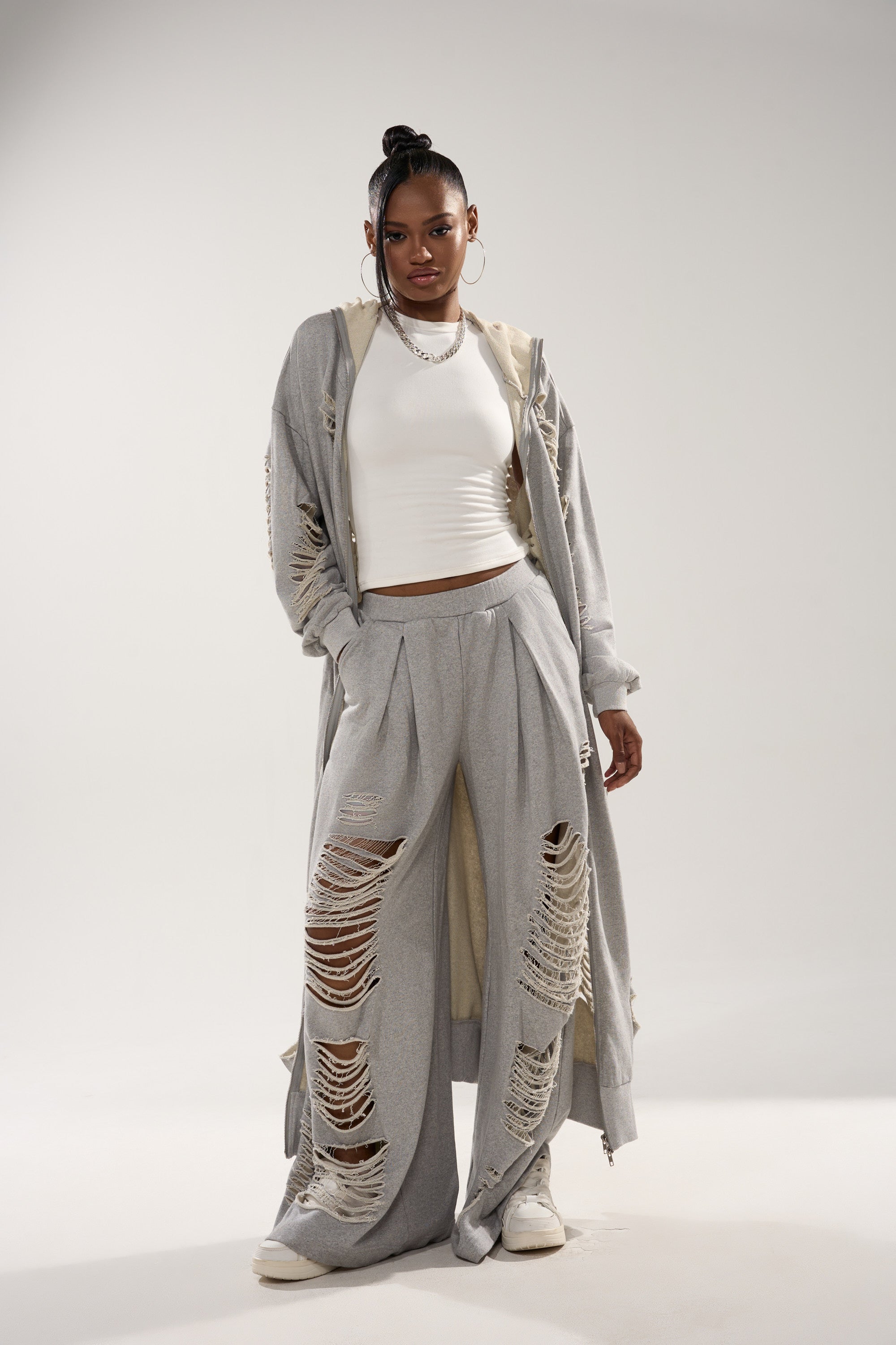 A woman stands confidently in the STRESS HIM OUT LONGLINE SWEATSHIRT—gray, distressed, and long—paired with matching ripped wide-leg pants, a white crop top, sneakers, and layered necklaces against a plain light background. 3