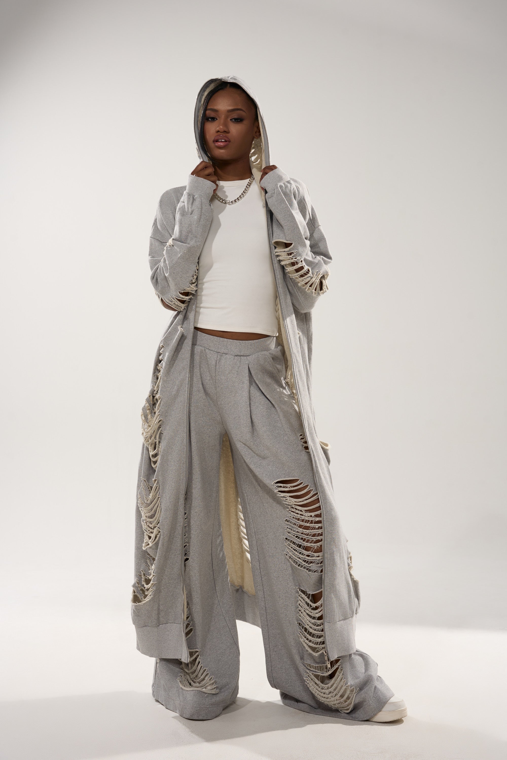 A woman poses confidently in the STRESS HIM OUT LONGLINE SWEATSHIRT, a light gray oversized hoodie coat with matching wide-leg pants and distressed loungewear details, styled with a white top and silver chain necklace against a plain background. 4