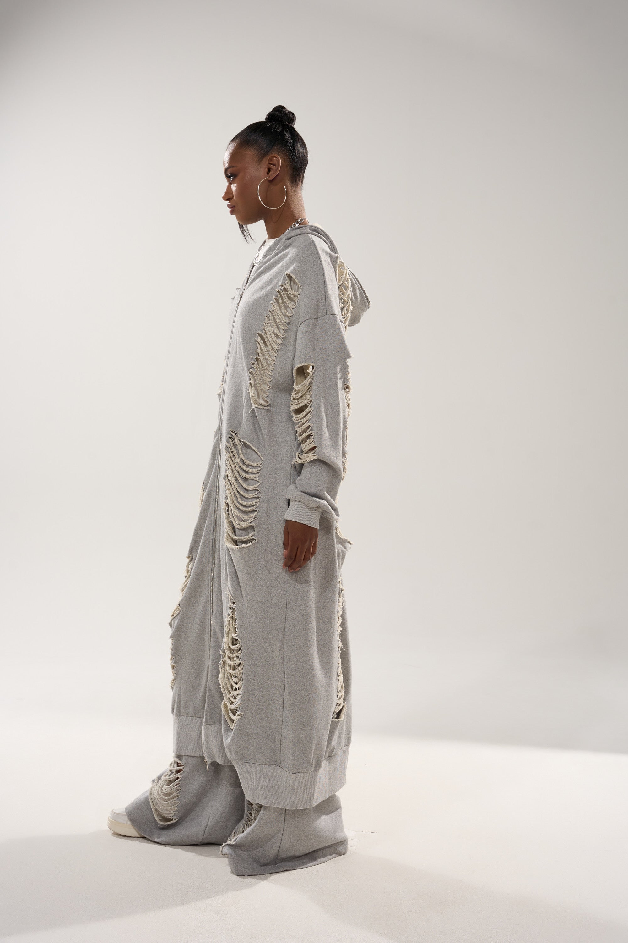 A person in profile wears the STRESS HIM OUT LONGLINE SWEATSHIRT, an oversized hoodie with large distressed holes, creating a textured, layered look, paired with wide-legged pants; their hair is styled in a bun. 5