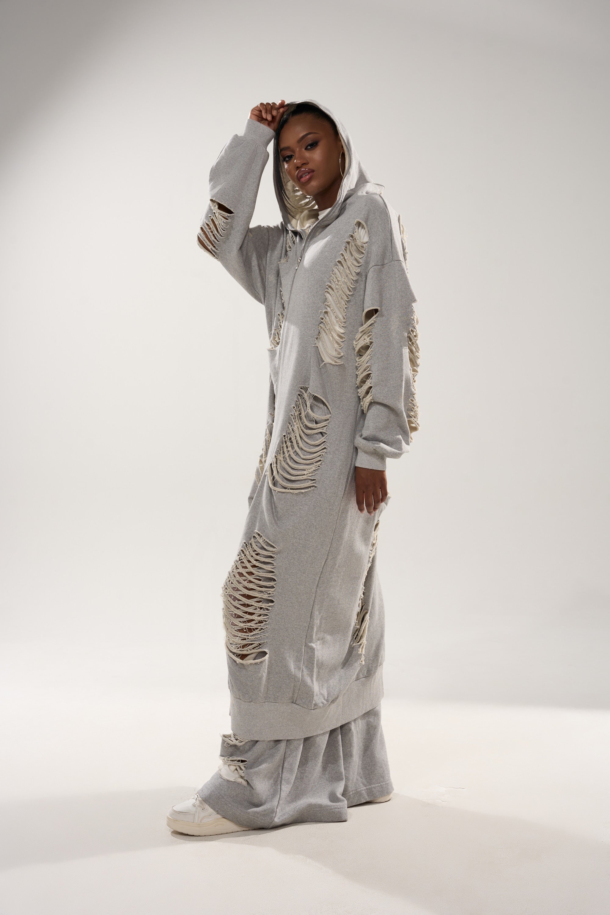 A person poses confidently in a studio wearing the STRESS HIM OUT LONGLINE SWEATSHIRT and ripped loungewear pants, with one hand lifting their hood against a plain, light background. 6