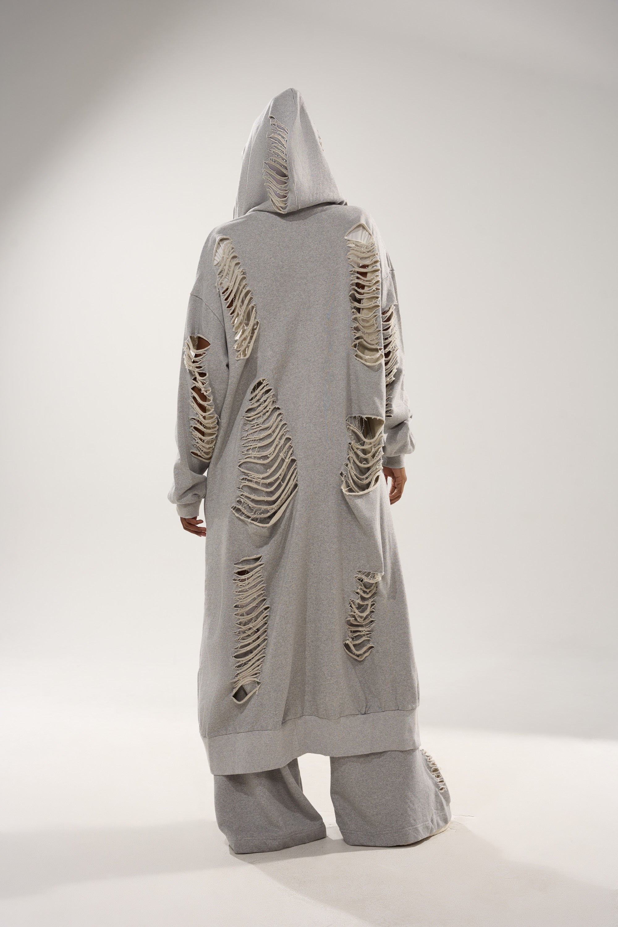 A person faces away in the STRESS HIM OUT LONGLINE SWEATSHIRT and matching pants, both light gray with large distressed holes. The plain, light background highlights the edgy loungewear set. 7