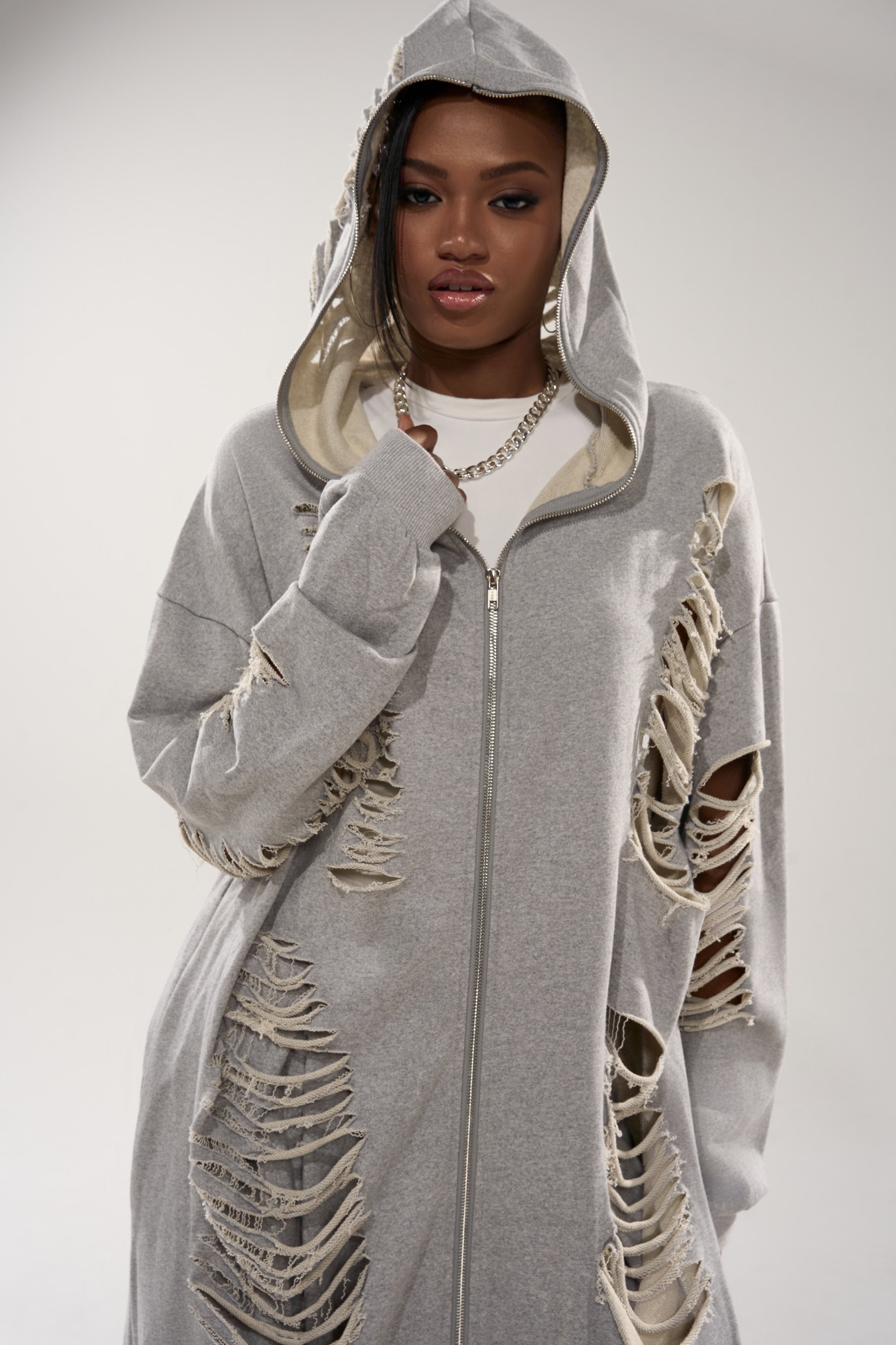 A woman faces the camera in the STRESS HIM OUT LONGLINE SWEATSHIRT, a grey, oversized hooded top with ripped details. Paired with a silver chain and a serious look, the light background highlights her modern loungewear style. 8