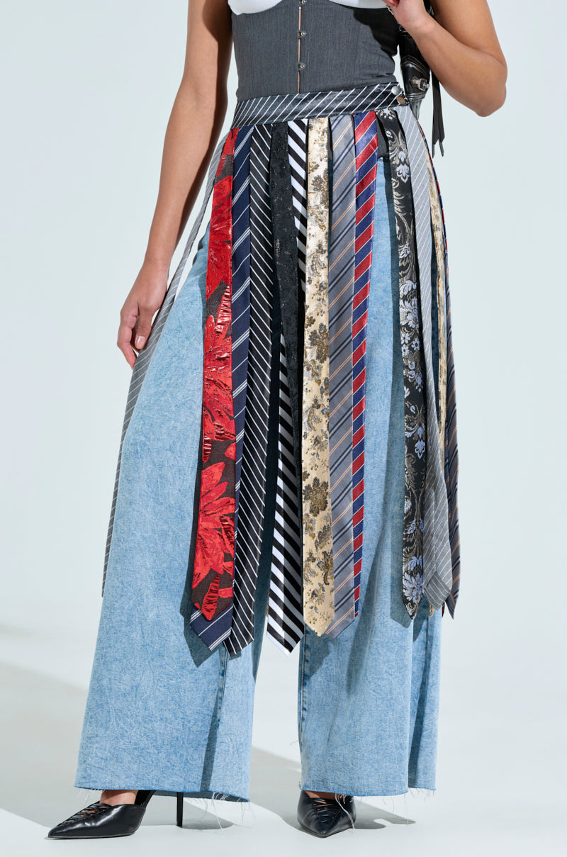 A fashion-forward person pairs wide-leg denim pants with the TIED IN BUSINESS MIDI SKIRT, a unique piece crafted from various neckties. Completing the look are a dark corset-style top, black pointed shoes, and metallic snap button closures for added flair.