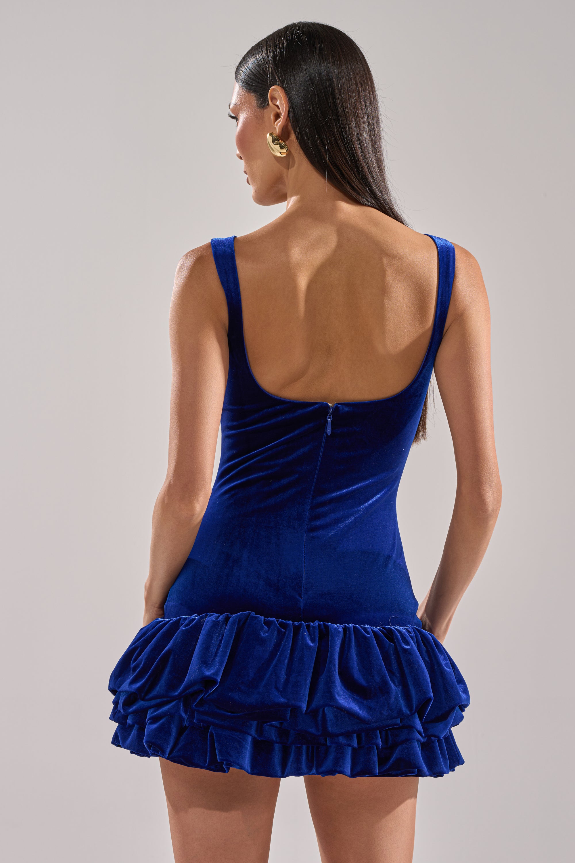 A woman with long, straight dark hair models the GIRL ON FIRE VELVET MINI DRESS IN BLUE, which features a low scoop back and layered ruffle skirt, standing against a plain light background.