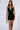 A person with long hair is wearing the CALL ME MESH MINI DRESS, which features a short black design with a plunging neckline and asymmetric strap detail. They are holding a silver handbag and standing against a plain background.