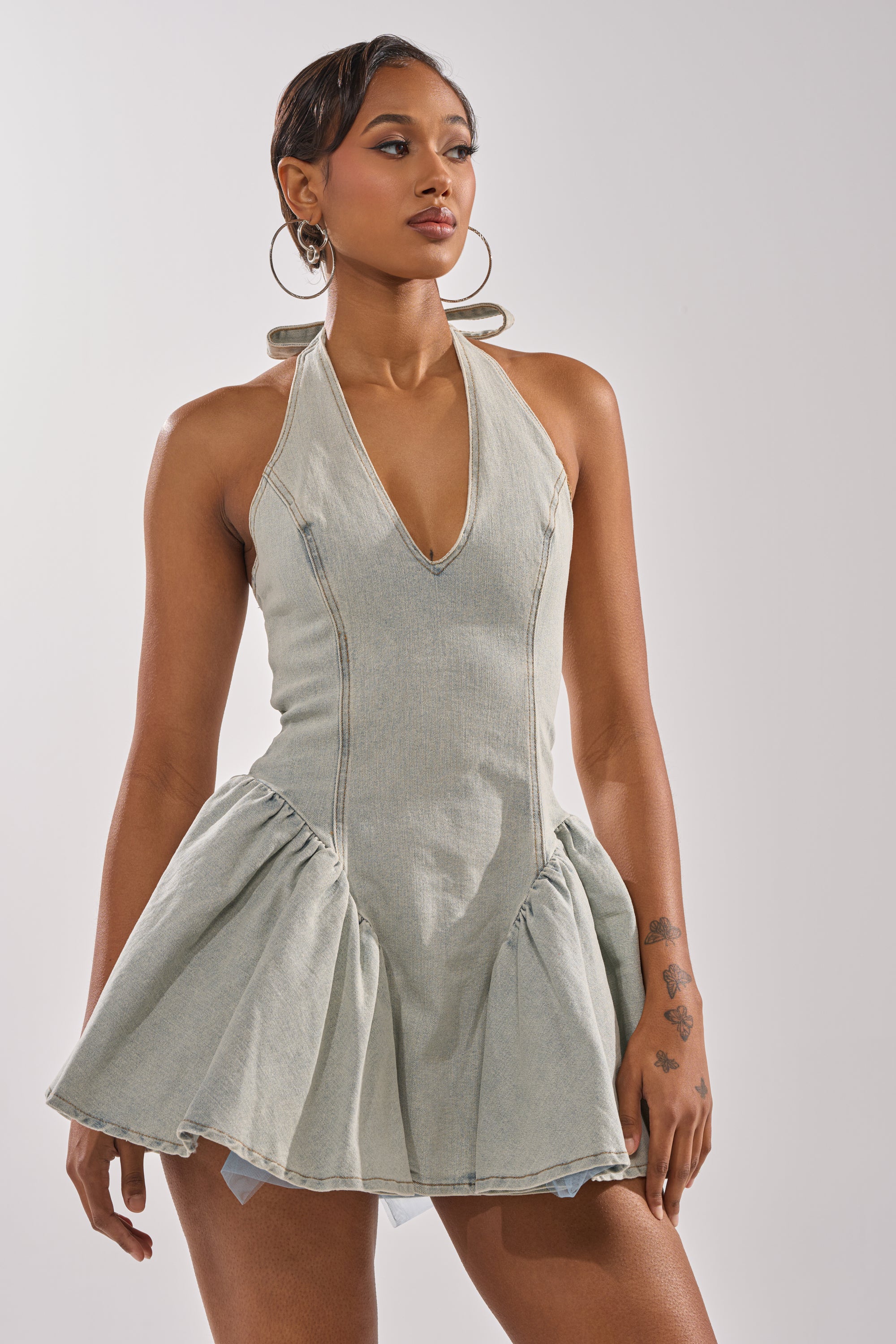A woman models the STYLE AND GRADE DENIM MINI DRESS, featuring a skater silhouette and flared skirt. She stands confidently against a light background, accessorized with hoop earrings and showcasing hand tattoos.