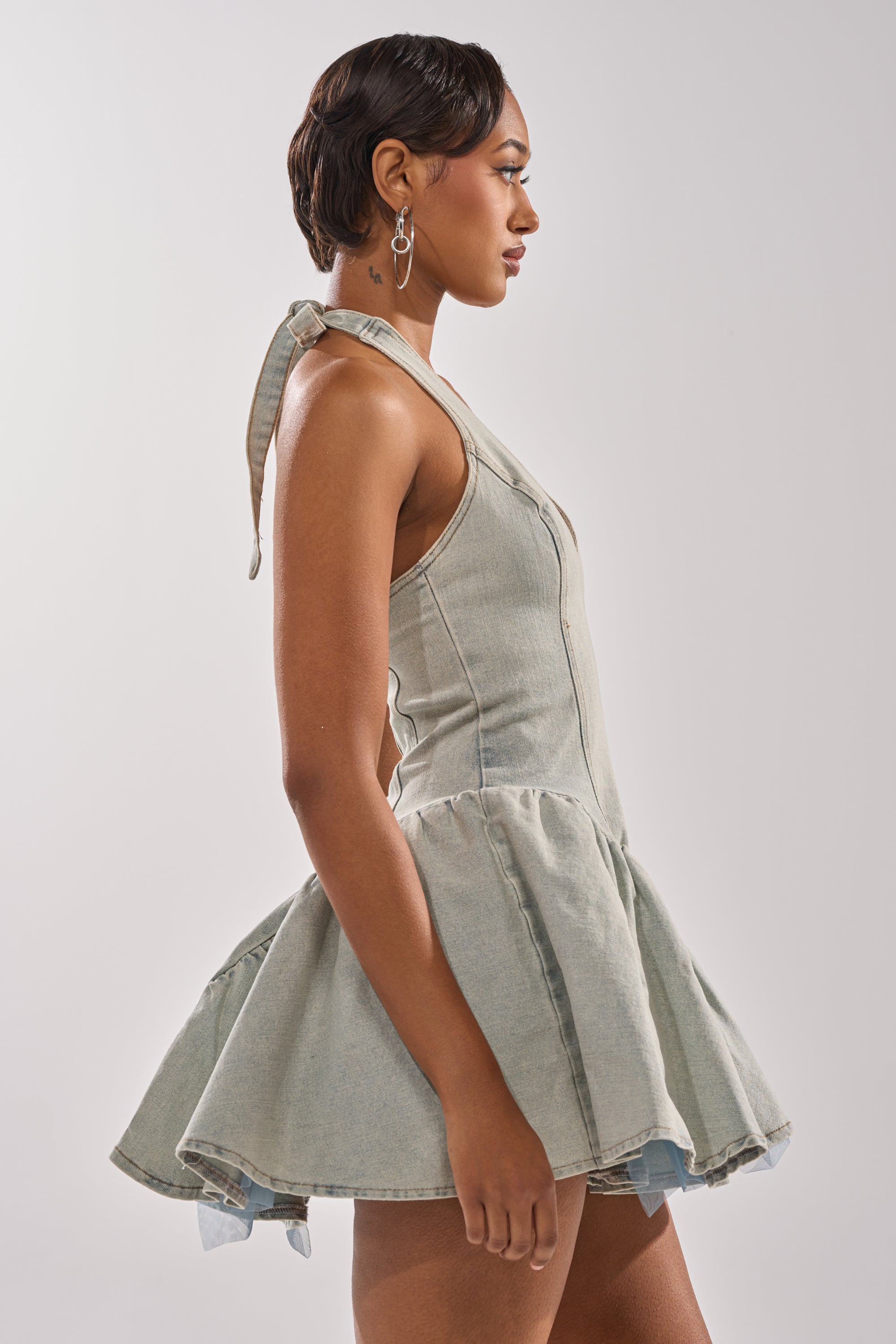 A woman with short dark hair and hoop earrings stands in profile against a plain background, wearing the STYLE AND GRADE DENIM MINI DRESS—a sleeveless mini dress with a skater silhouette and flared skirt.