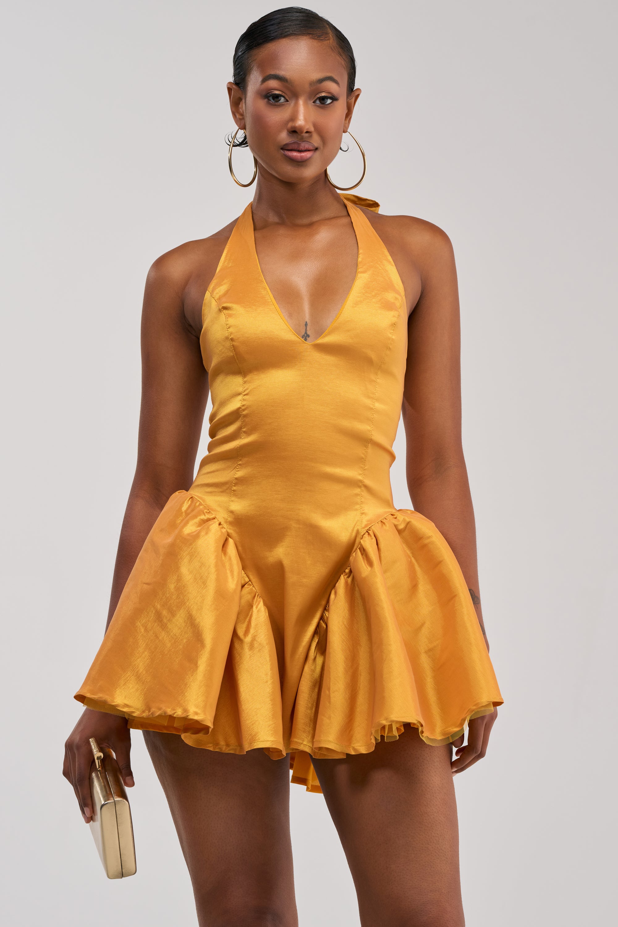 A woman stands against a plain light background wearing the STYLE AND GRACE MINI DRESS IN MUSTARD TAFFETA—a sleeveless, halter mini dress with a fitted bodice and flared skirt—evoking an elegant homecoming look, while holding a small cream clutch.