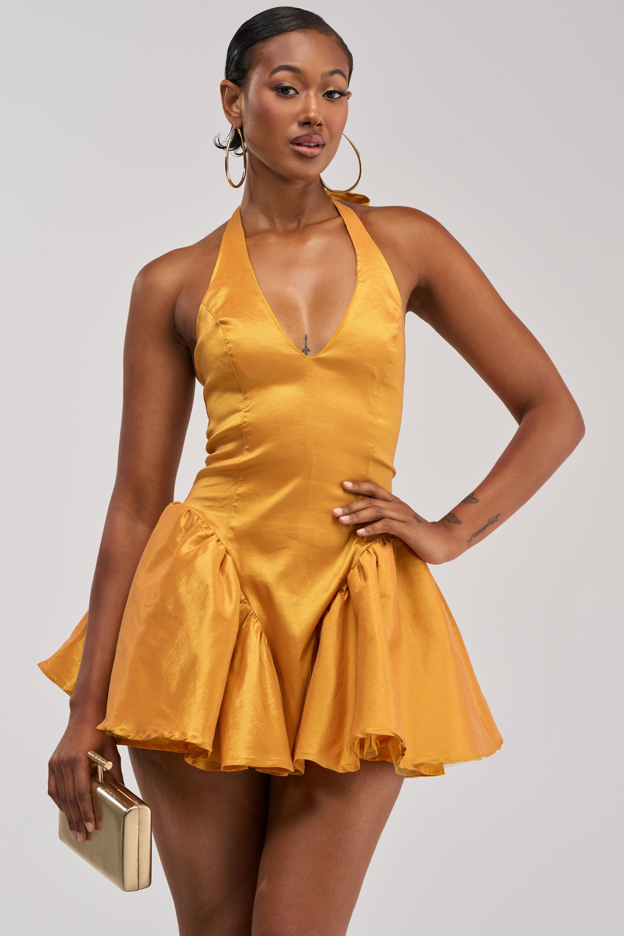 A woman poses confidently against a plain background wearing the STYLE AND GRACE MINI DRESS IN MUSTARD TAFFETA, featuring a sleeveless design, deep V-neckline, and flared mini skirt. She accessorizes with hoop earrings and holds a beige clutch.