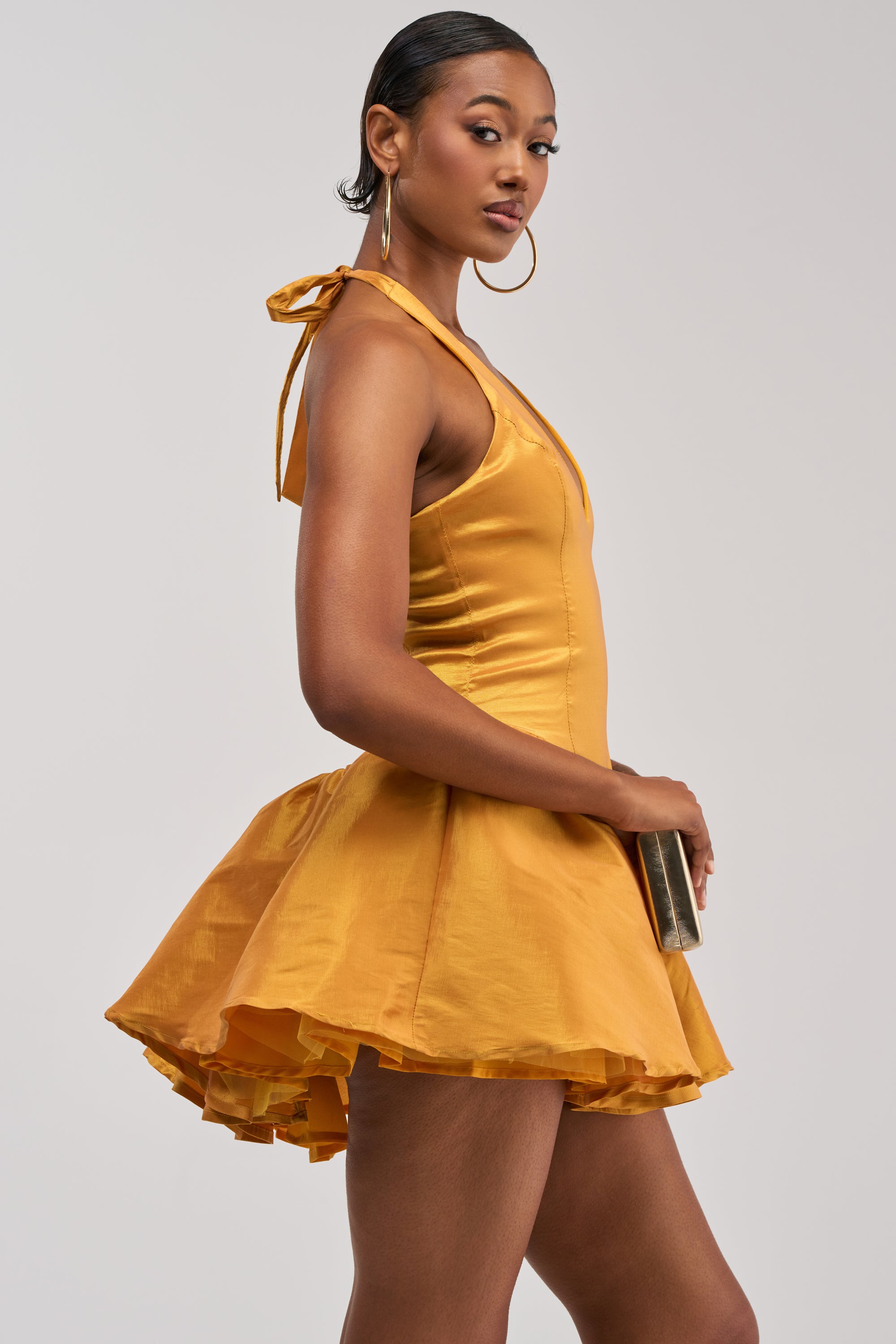 Wearing the STYLE AND GRACE MINI DRESS IN MUSTARD TAFFETA—with its shiny fabric, mustard yellow hue, and flared mini skirt—a woman stands in profile against a plain, light background while holding a rectangular clutch.