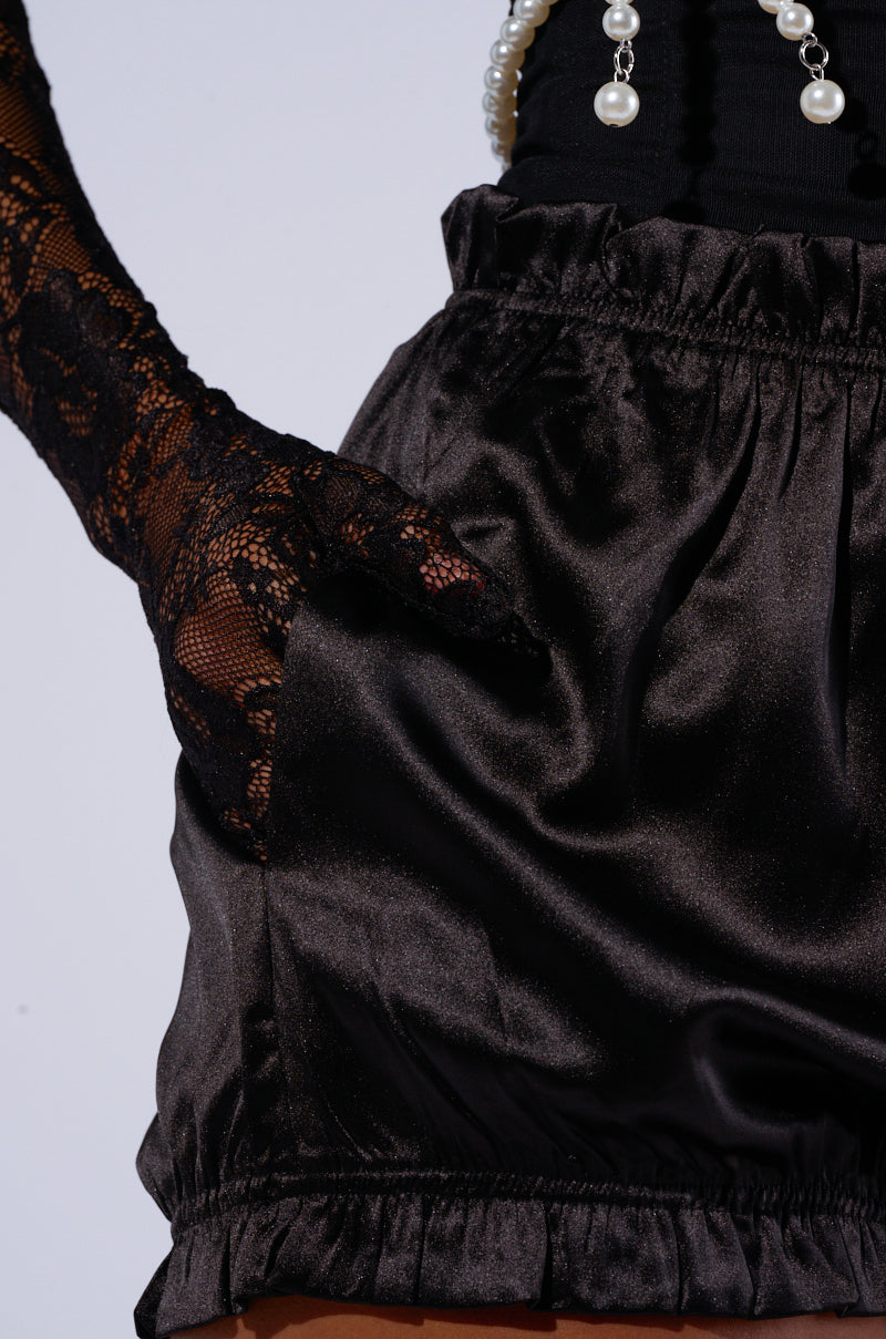 A person is wearing the BETSY BLOOMER SHORT, featuring a high-rise fit in black satin with a ruffled waistband and a hand in the pocket. The ensemble includes an elegant long black lace glove and a stylish black top accessorized with a white pearl necklace. The setting is plain and uncluttered. 6