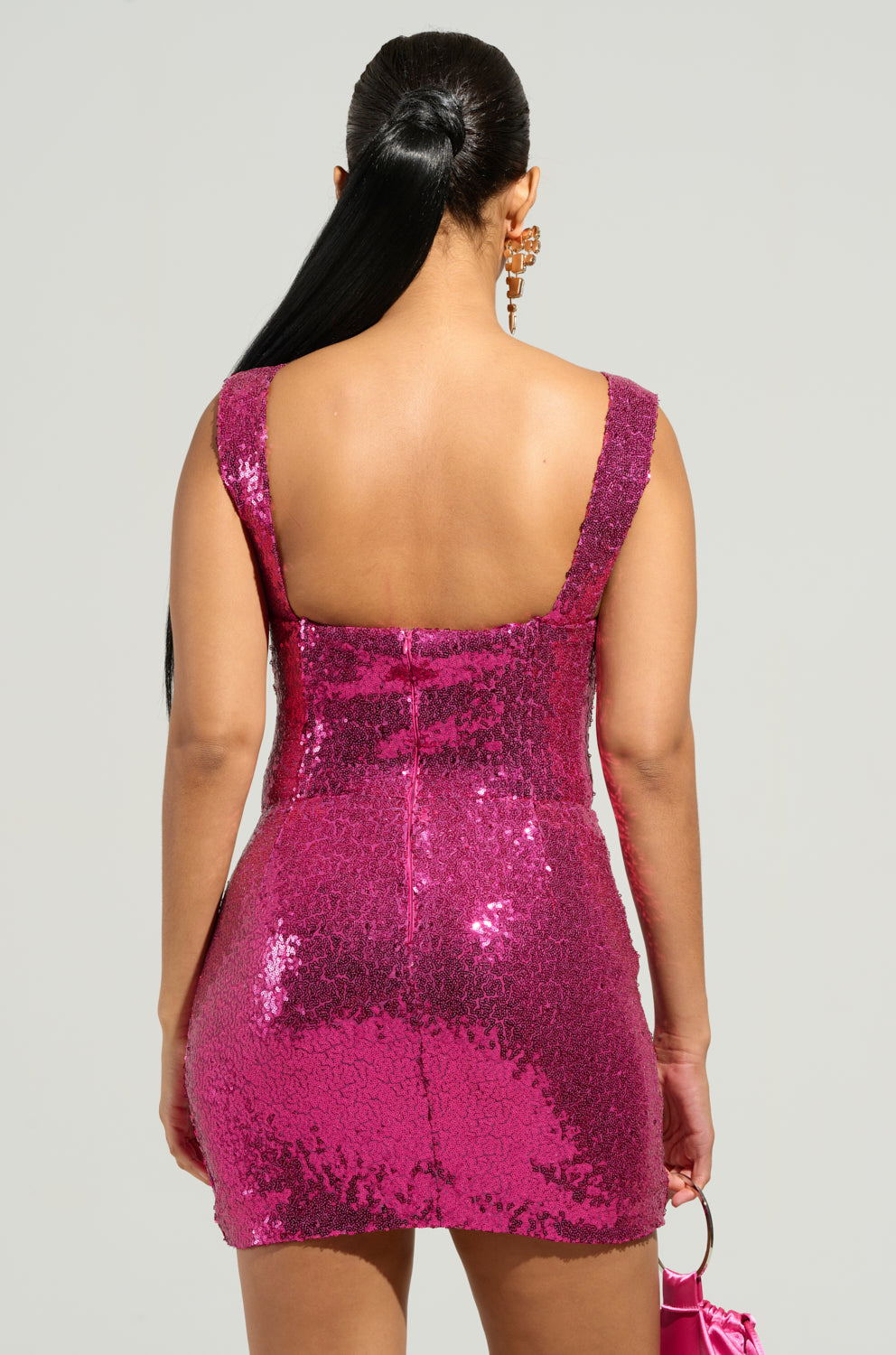 A woman with long black hair, wearing the DANCE ALL NIGHT SEQUIN CORSET MINI DRESS in shimmering pink, stands with her back to the camera. She holds a small pink handbag and wears large, gold hoop earrings. The bodycon silhouette of her outfit enhances her style against the plain, light-colored background. 2