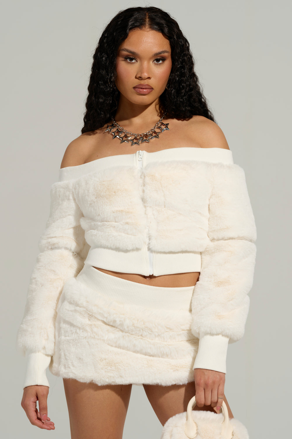 Wearing the COLDEST WINTER OFF THE SHOULDER FUR TOP IN WHITE, a woman with curly hair pairs it beautifully with a coordinating skirt. She adds flair with a chunky necklace and clutches a small faux sherpa bag. A neutral backdrop enhances her fashionable look.