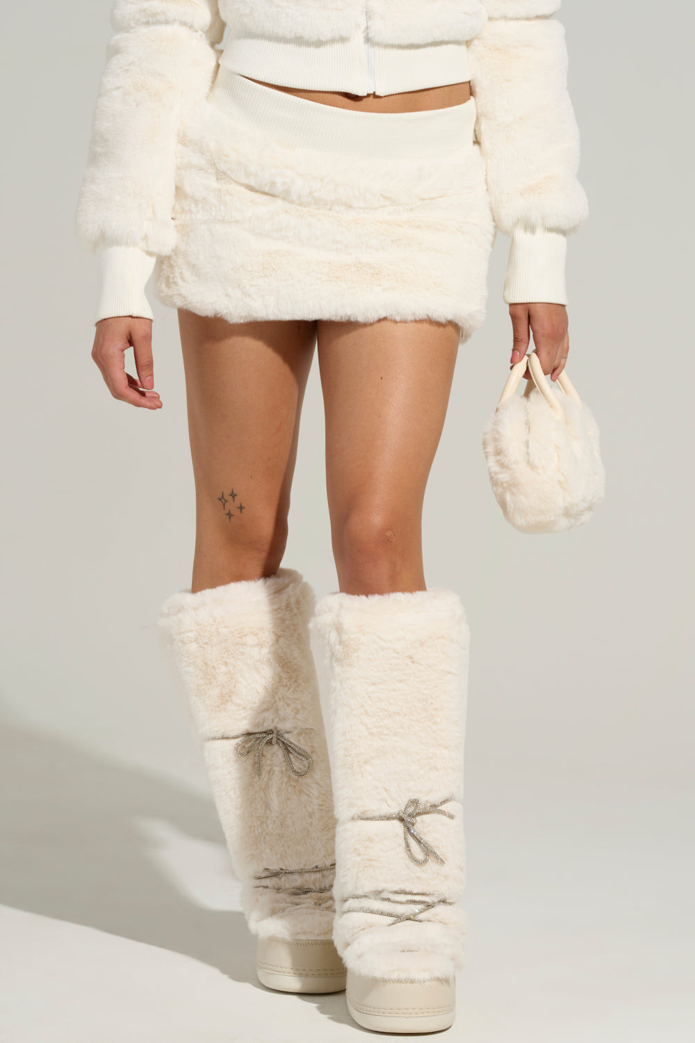 A person is dressed in the COLDEST WINTER FUR MINI SKIRT IN WHITE, featuring a fluffy texture and high-rise fit. They complement it with a matching jacket and large furry boots while holding a small coordinating furry bag. This minimalist ensemble is styled perfectly for chic cold-weather fashion.