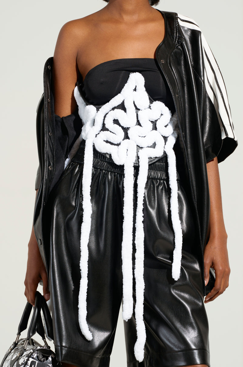 A person wearing a unique fashion outfit: a black faux leather jacket and shorts, a black top paired with the AESTHETIC APPLIQUE CORSET BELT, and a white knotted rope accessory draped over the torso. They carry a handbag with a patterned design.