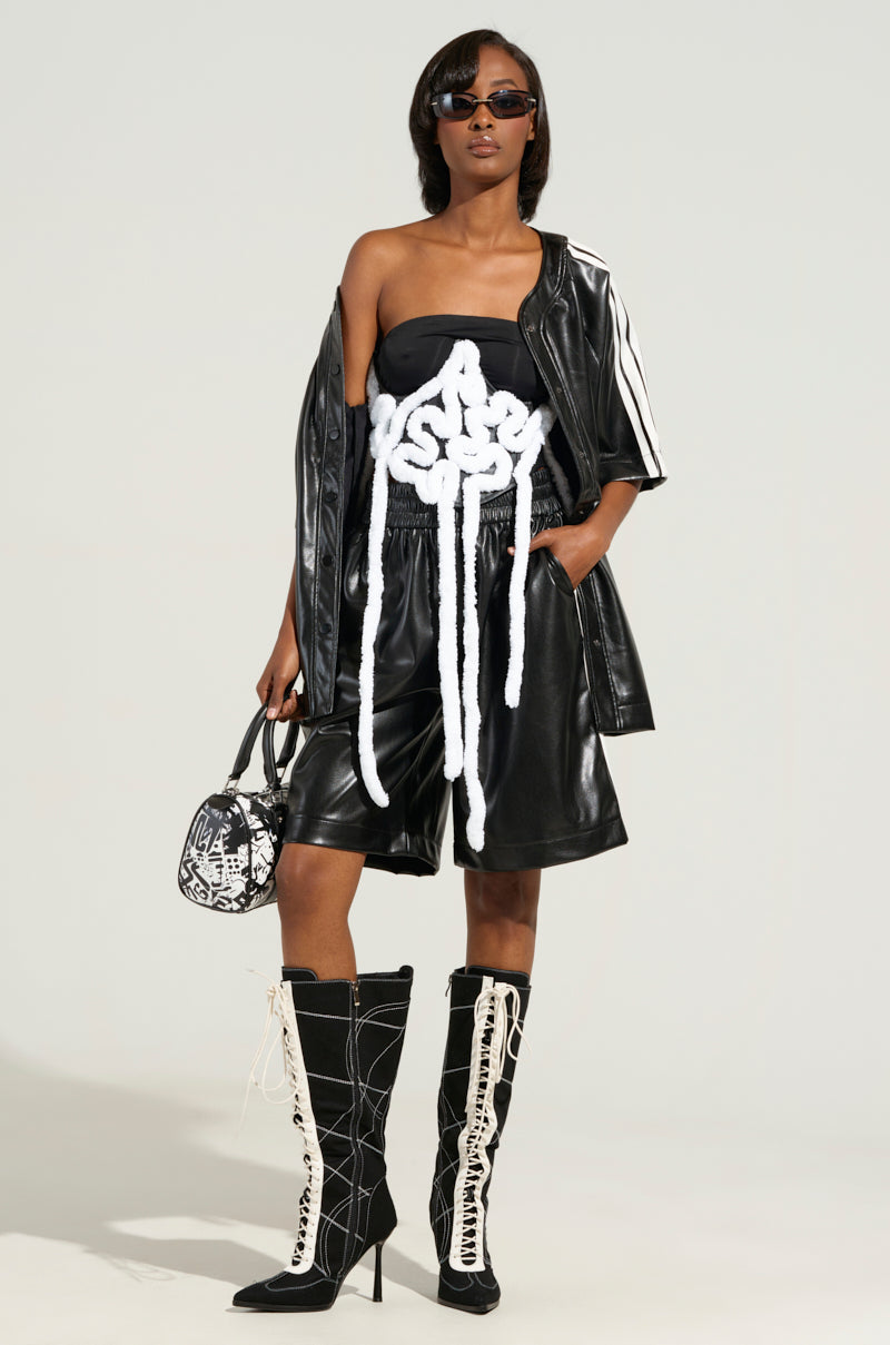 A person wearing sunglasses and a black and white outfit with a striking white design on top stands confidently against a plain background, their look enhanced by a faux leather jacket. The ensemble is completed with knee-high lace-up boots, the AESTHETIC APPLIQUE CORSET BELT accentuating the outfit, and a small patterned bag.