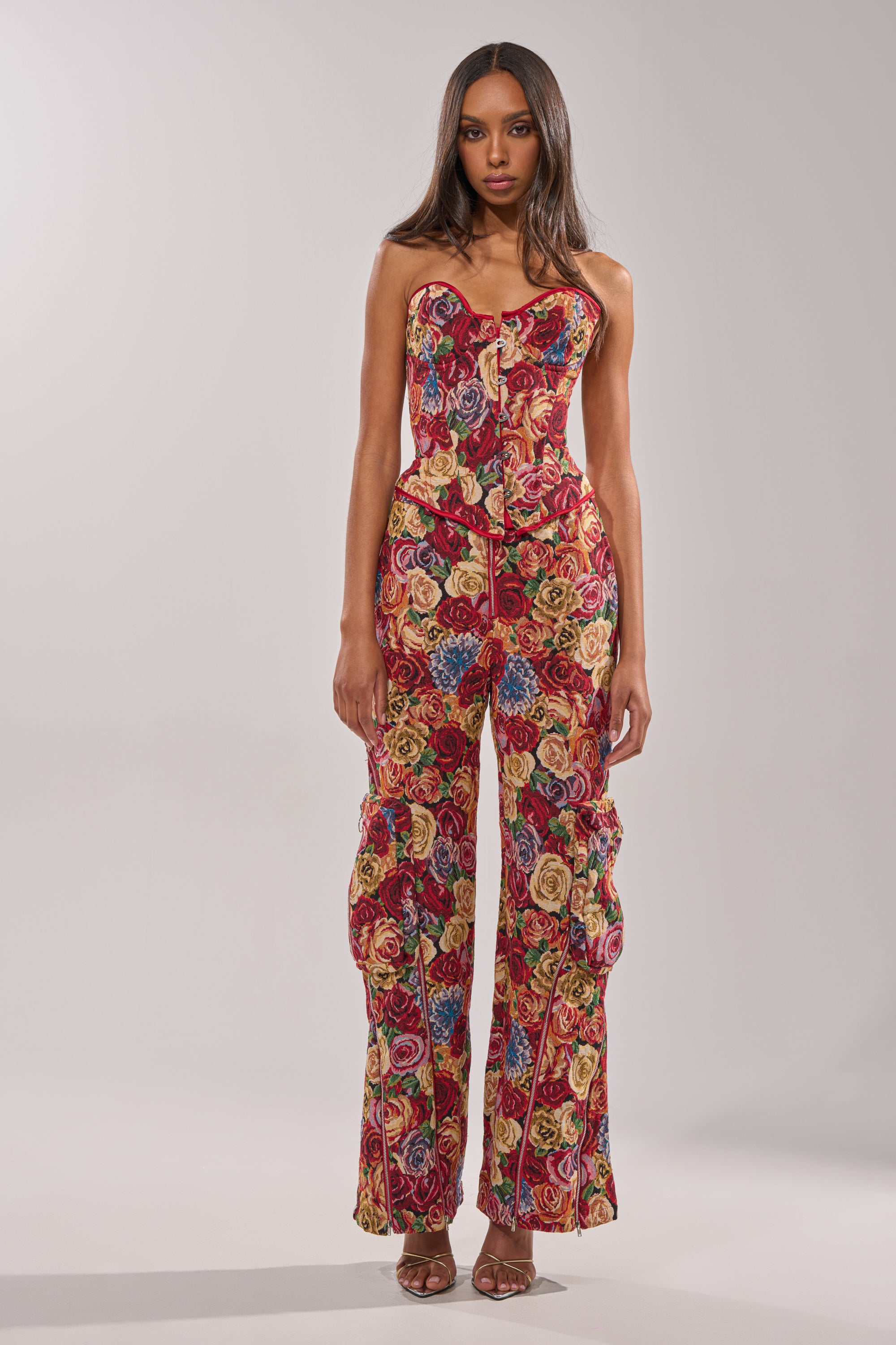 A woman models the INTO THE THICK OF IT FLORAL BROCADE CORSET, a strapless piece with vibrant red, yellow, and blue floral patterns. She pairs it with brown heeled sandals and has long, straight hair against a plain light background. 6