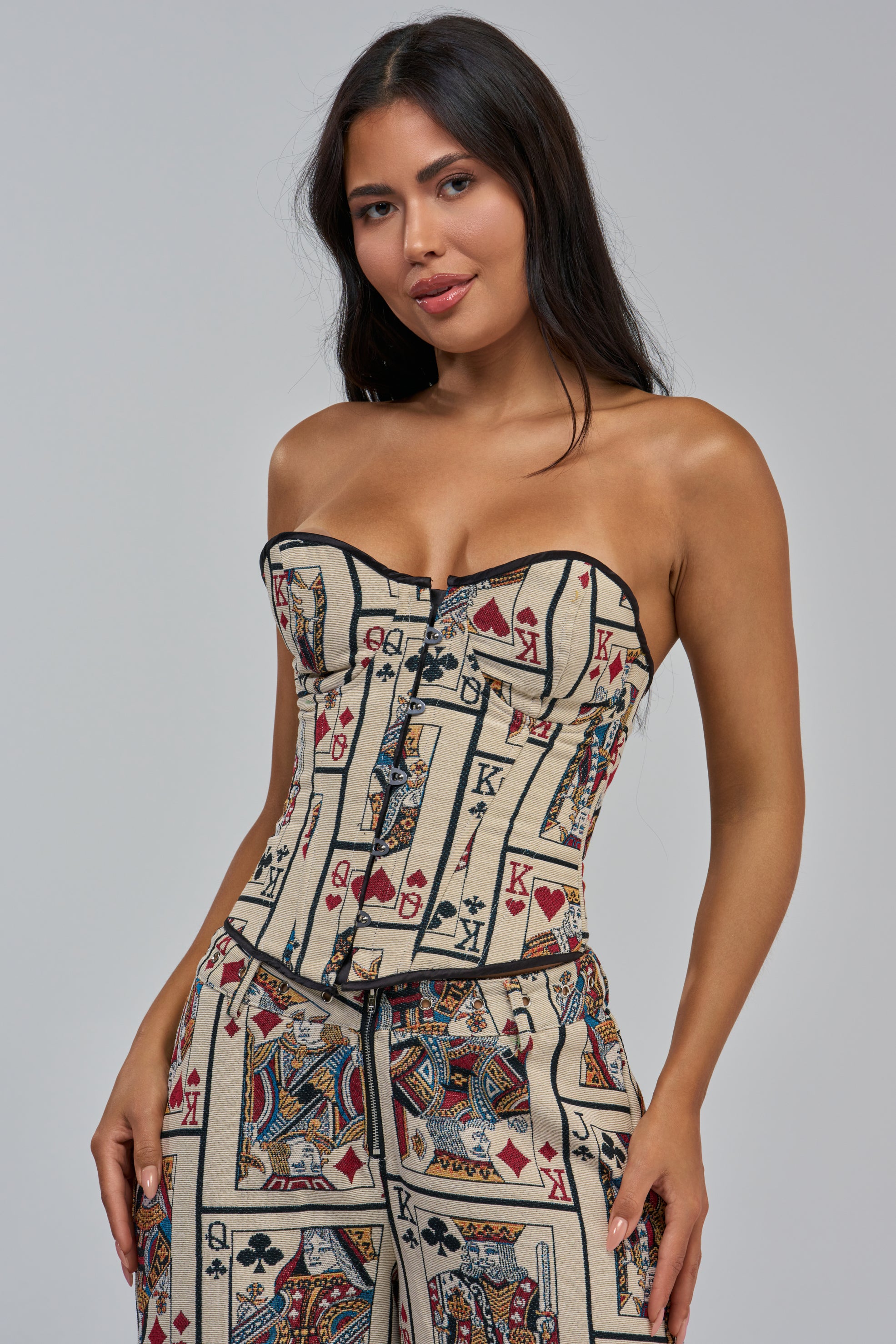 A woman wears the WIN ME SOME MONEY CARD CORSET top and matching pants, both showcasing a bold playing card print with king, queen, and ace motifs in red, black, and gold on a white jacquard brocade background, standing against a plain grey backdrop.