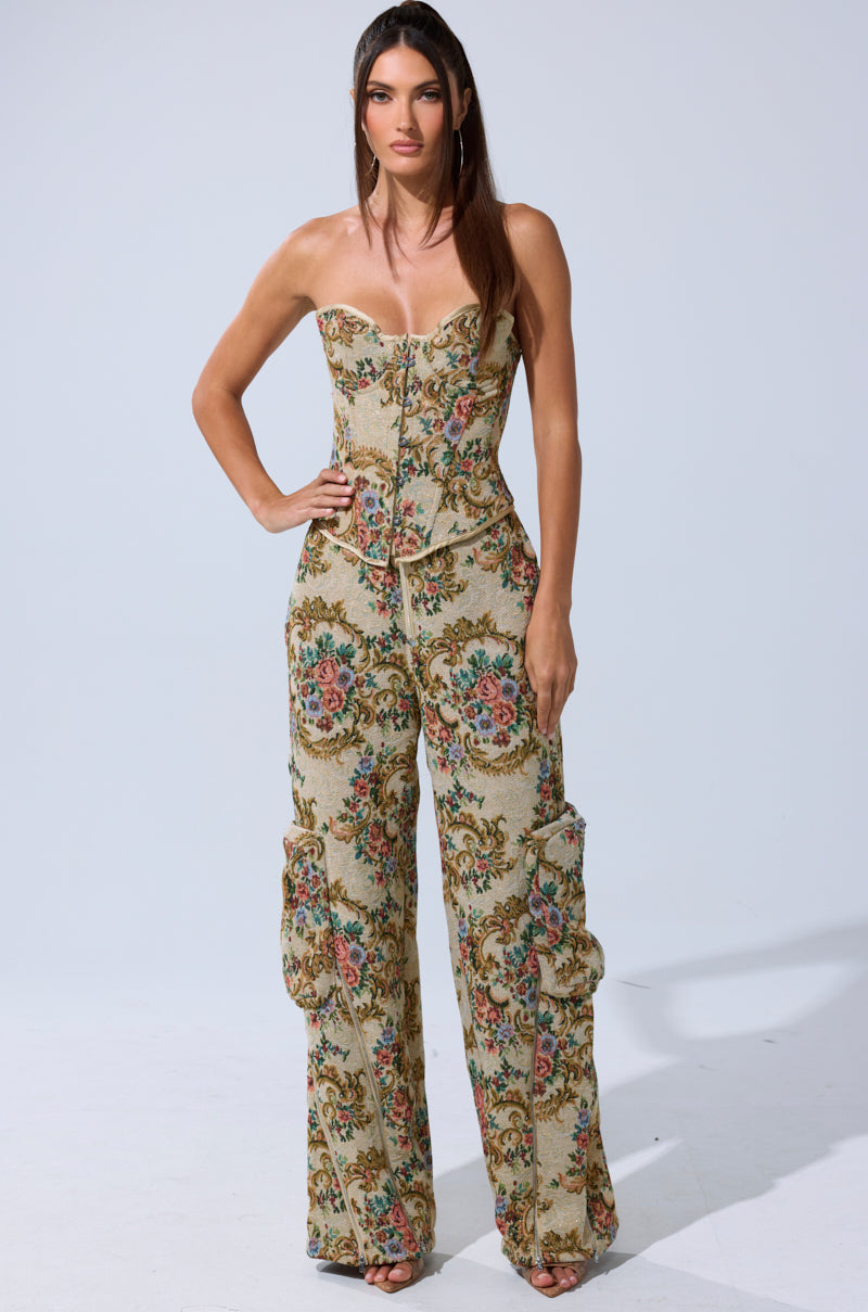 A woman stands against a plain background wearing the SHINE AWAY BROCADE TROUSER—high-rise, wide-leg pants with ruffle details and a vintage-style tapestry floral pattern with metallic gold accents, paired with a matching fitted corset top.