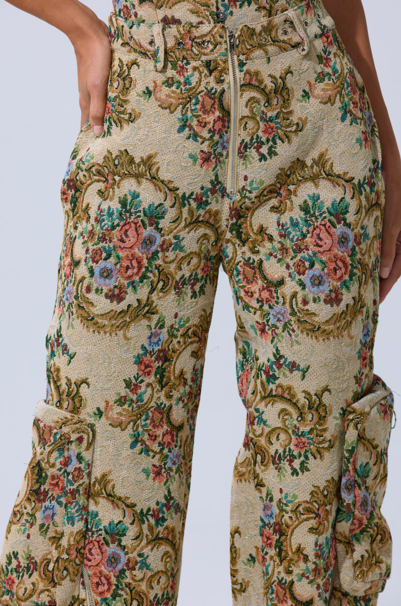 A close-up shows a person wearing the SHINE AWAY BROCADE TROUSER, featuring a high waist, vintage floral tapestry in green, pink, red, blue, and beige tones. One hand rests on their hip; the wide-leg silhouette creates a statement look.