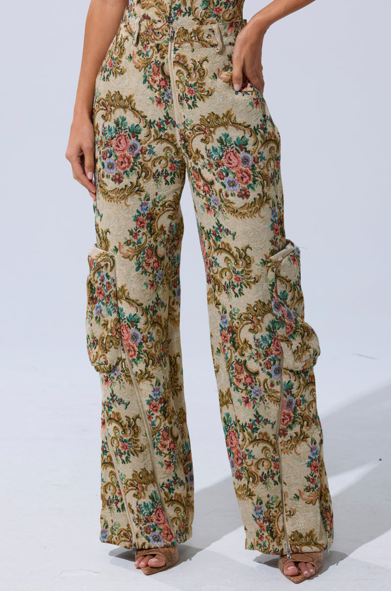 Wearing the SHINE AWAY BROCADE TROUSER, featuring a wide-leg silhouette and ornate floral tapestry in gold, pink, green, and blue with visible seams and pockets, they pose against a plain light background.