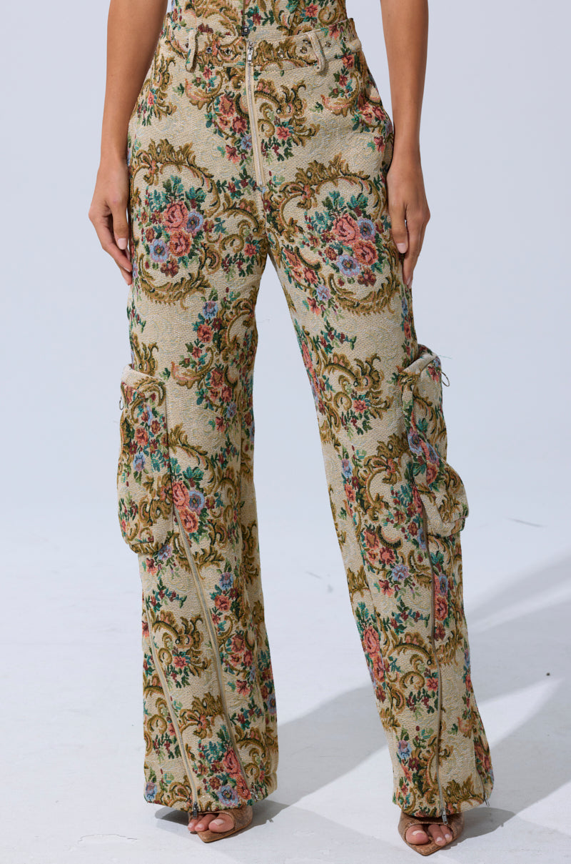 Wearing the SHINE AWAY BROCADE TROUSER—high-waisted, wide-leg pants with an ornate green, pink, and gold floral scrollwork tapestry—someone stands against a plain light background.