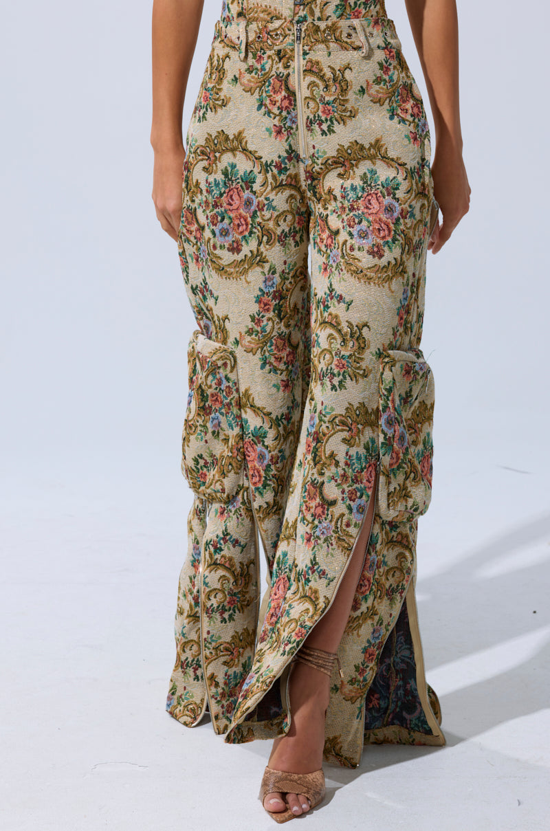 Someone wears the SHINE AWAY BROCADE TROUSER—high-waisted, wide-leg pants with a vintage floral tapestry pattern, front slits, and large side pockets—styled with open-toe heeled sandals.