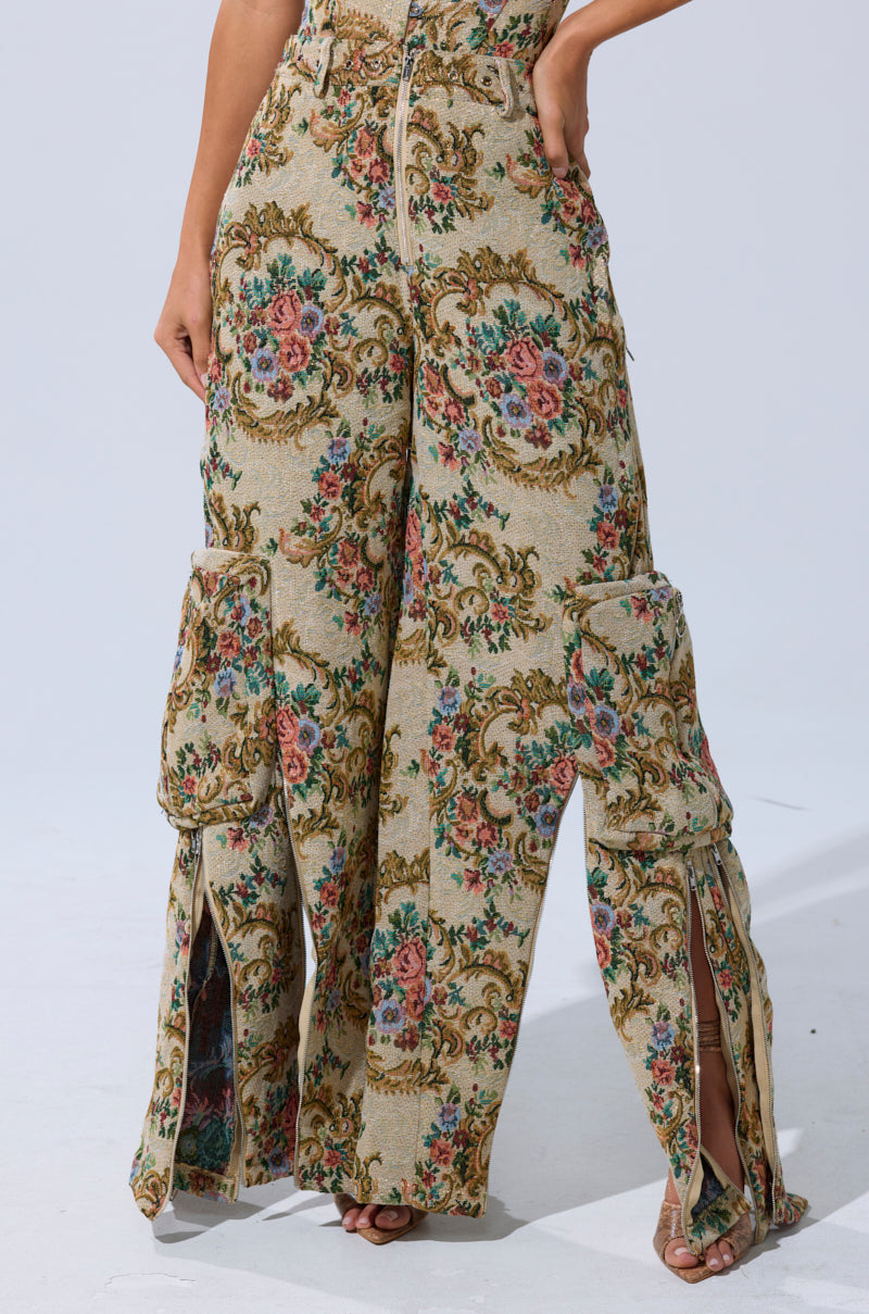 Wearing the SHINE AWAY BROCADE TROUSER, featuring a vintage floral tapestry pattern, wide legs, cargo pockets, and front slits, with their hand on their hip and only their lower half visible.