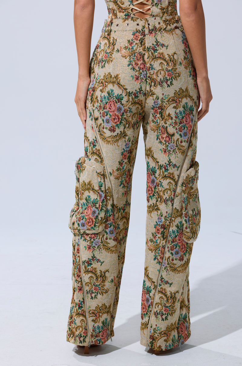Someone wearing the SHINE AWAY BROCADE TROUSER—high-waisted, floral-patterned, wide-leg cargo pants with side pockets—stands with their back to the camera against a plain light background.