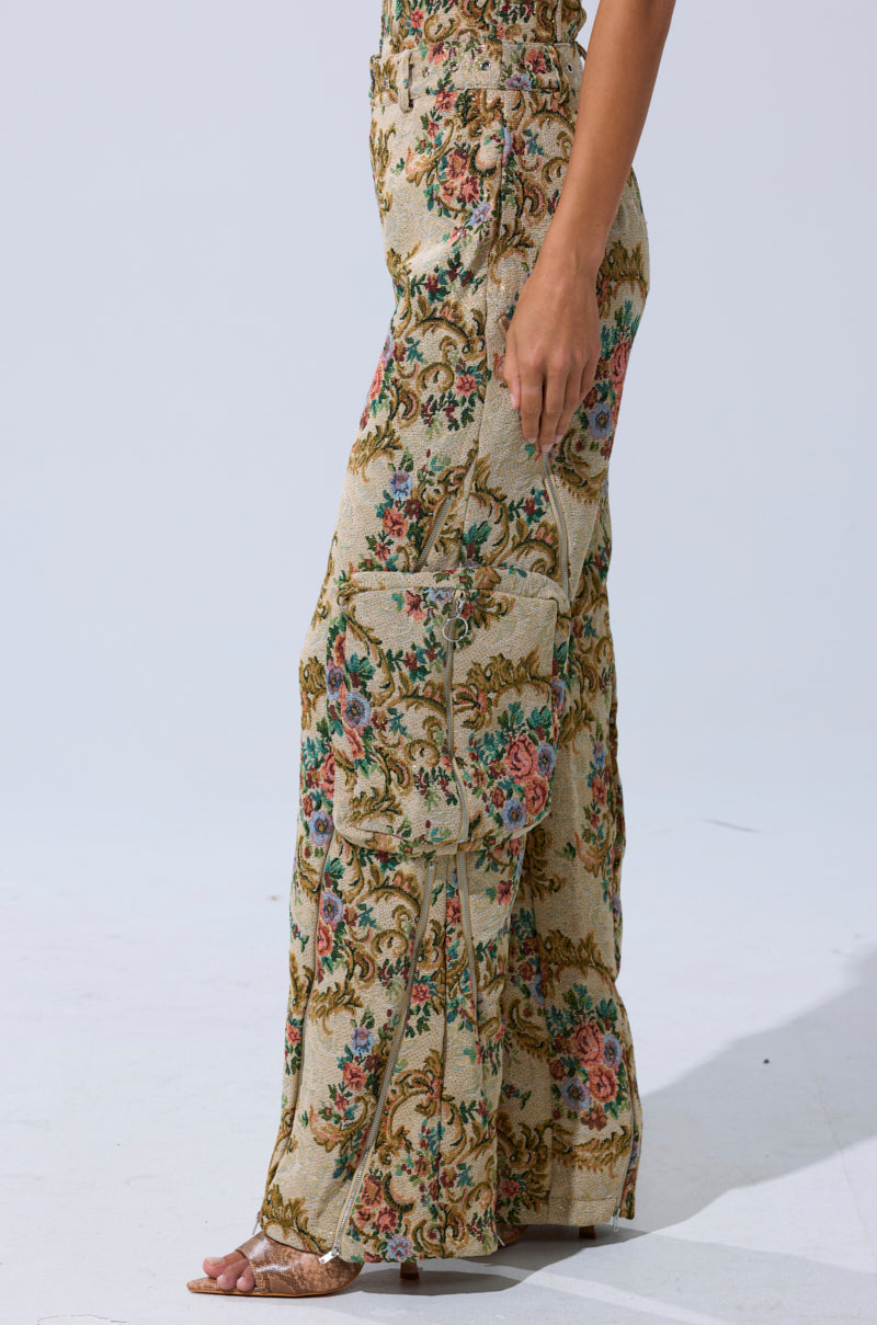 Model in high-waisted, wide-leg trousers featuring an ornate floral tapestry pattern and large side pocket, shown from the side against a plain light background, showcases the SHINE AWAY BROCADE TROUSER.