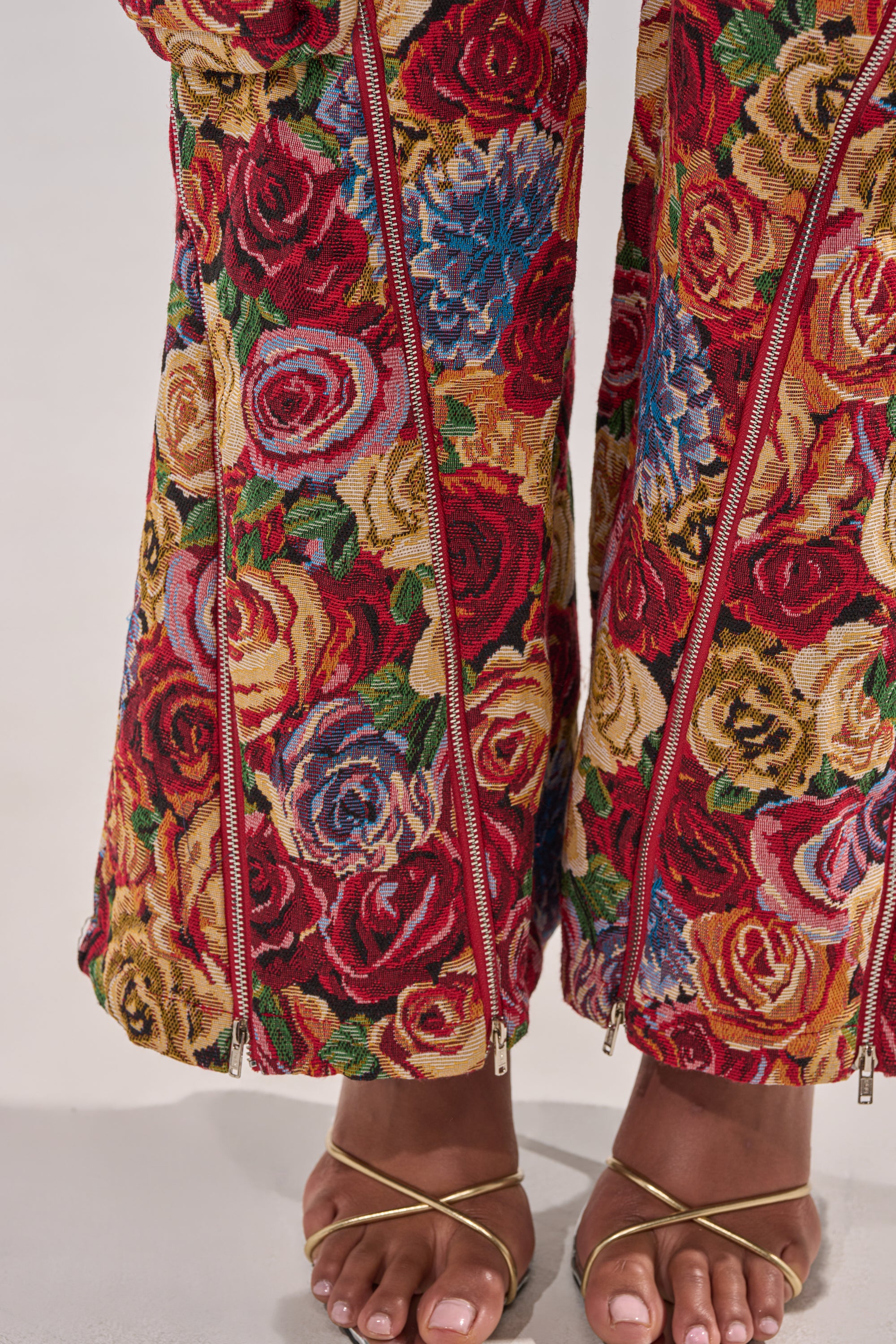 A close-up of a person wearing INTO THE THICK OF IT FLORAL CARGO PANT with vintage rose print and gold zippers, paired with gold strappy sandals showing their toes on a light background. 9
