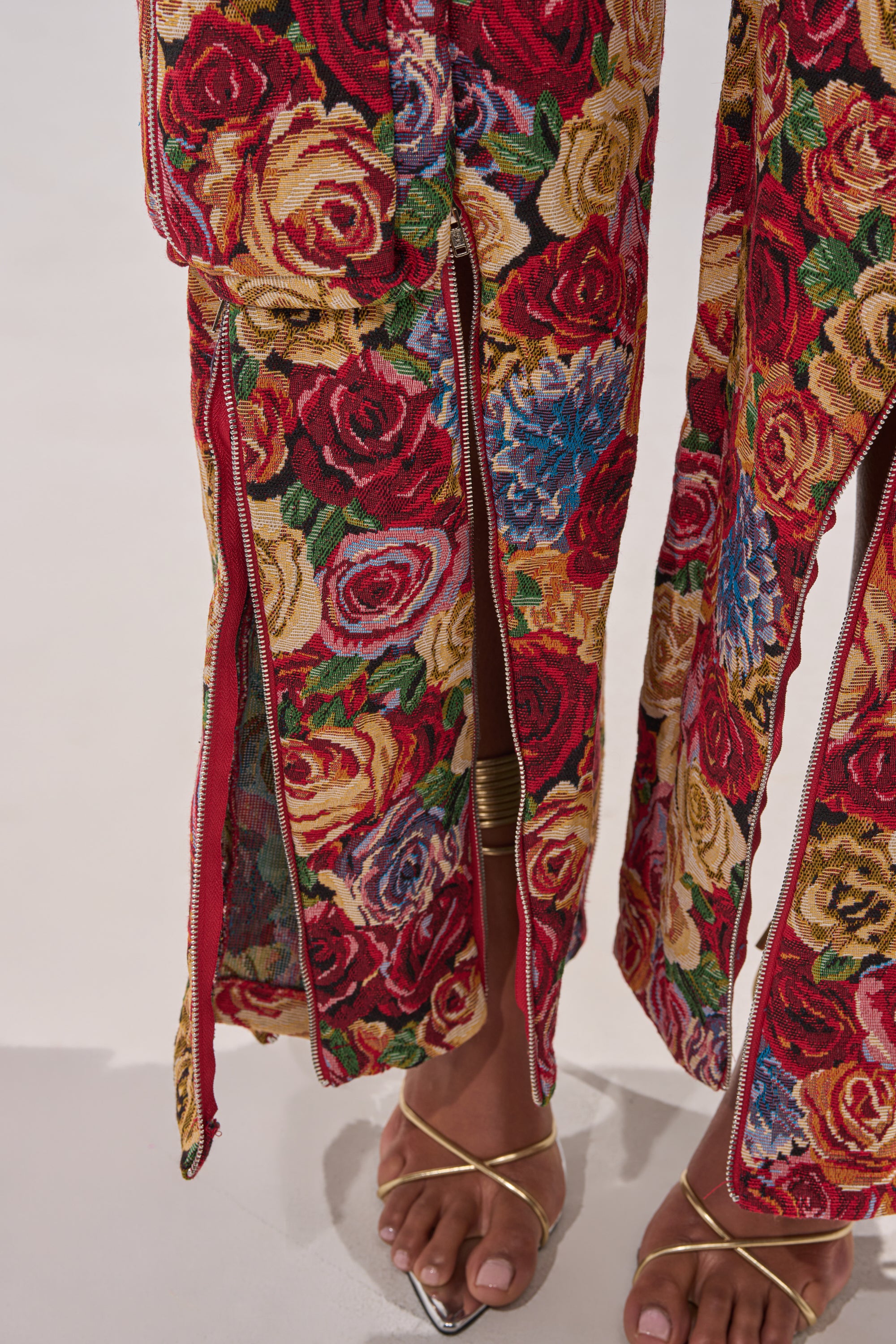 Close-up of a person wearing the INTO THE THICK OF IT FLORAL CARGO PANT featuring a vibrant floral print, wide legs with zippered slits at the hem, styled with gold strappy sandals and a gold ankle cuff. 10