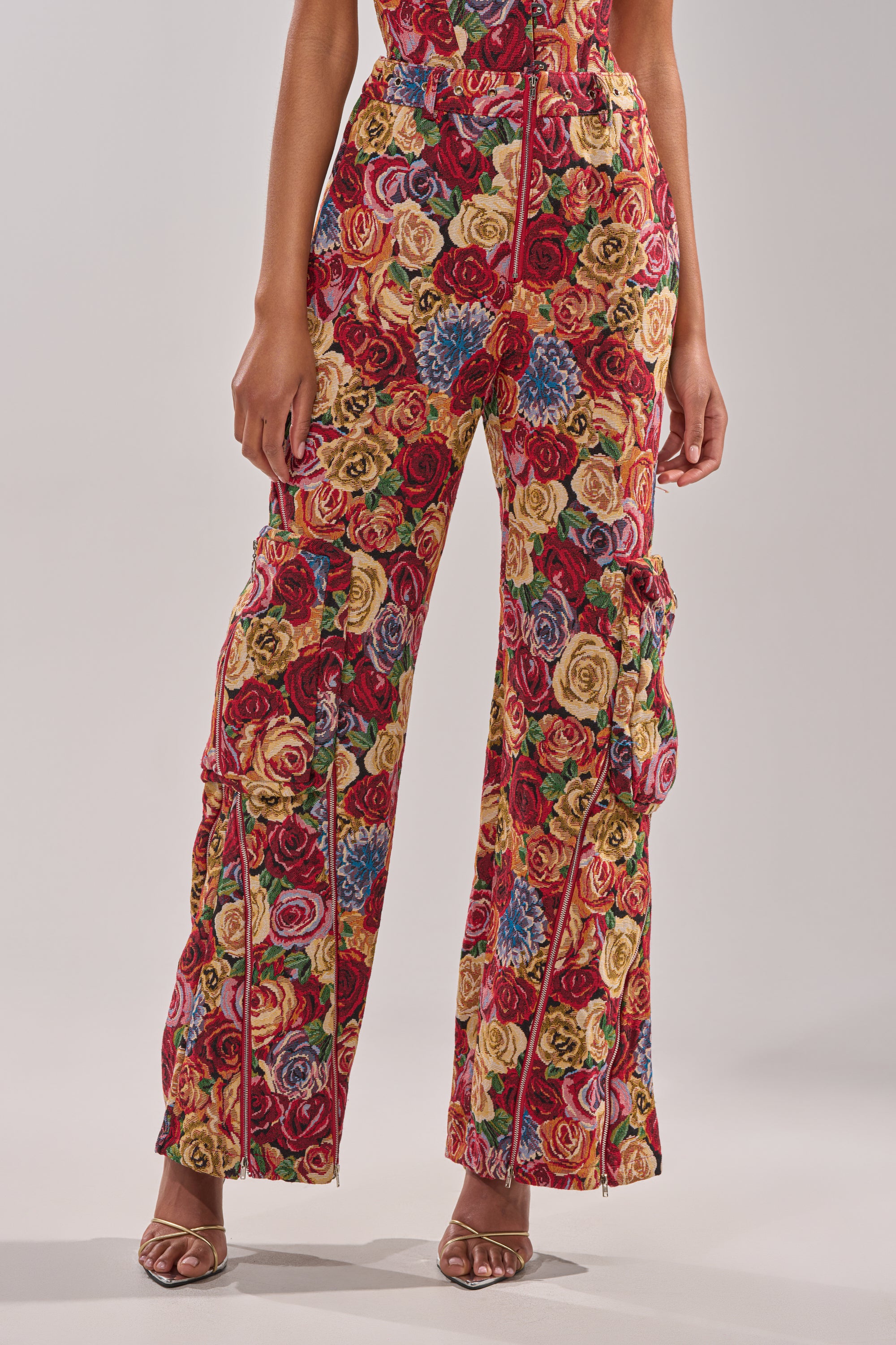 A person models the INTO THE THICK OF IT FLORAL CARGO PANT, featuring a vibrant vintage rose print in red, yellow, and blue. Only their lower body in heeled sandals is visible against a plain light background. 1