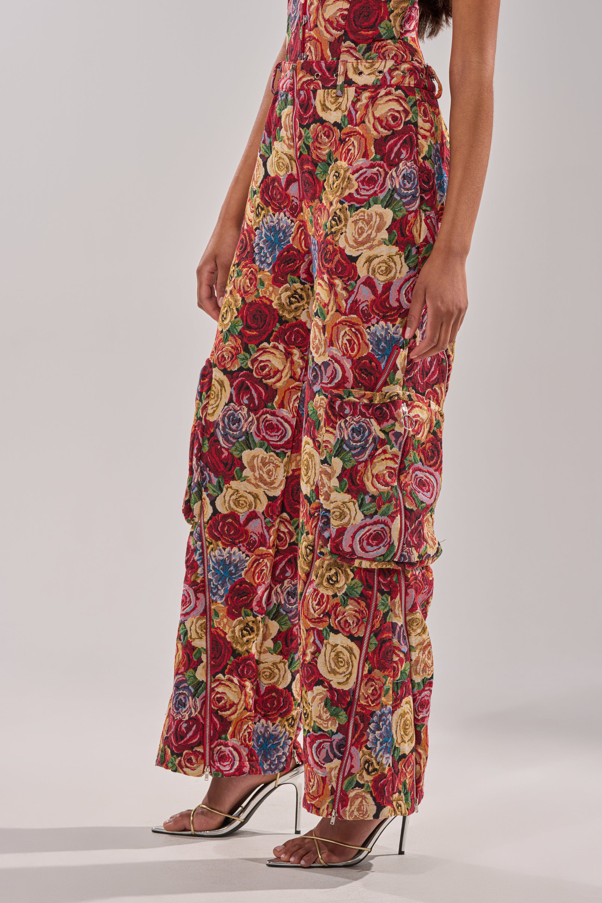 Model wears the INTO THE THICK OF IT FLORAL CARGO PANT featuring vibrant floral patterns and large pockets, paired with silver high-heeled sandals, shown from shoulder to foot against a plain background. 2