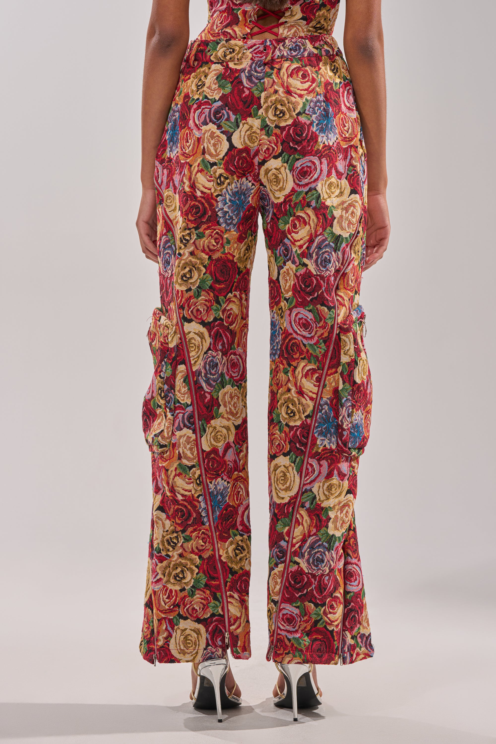 A person stands facing away, wearing the INTO THE THICK OF IT FLORAL CARGO PANT—high-waisted, flared pants with a floral tapestry in red, yellow, blue, and green tones—paired with white high-heeled sandals. 3