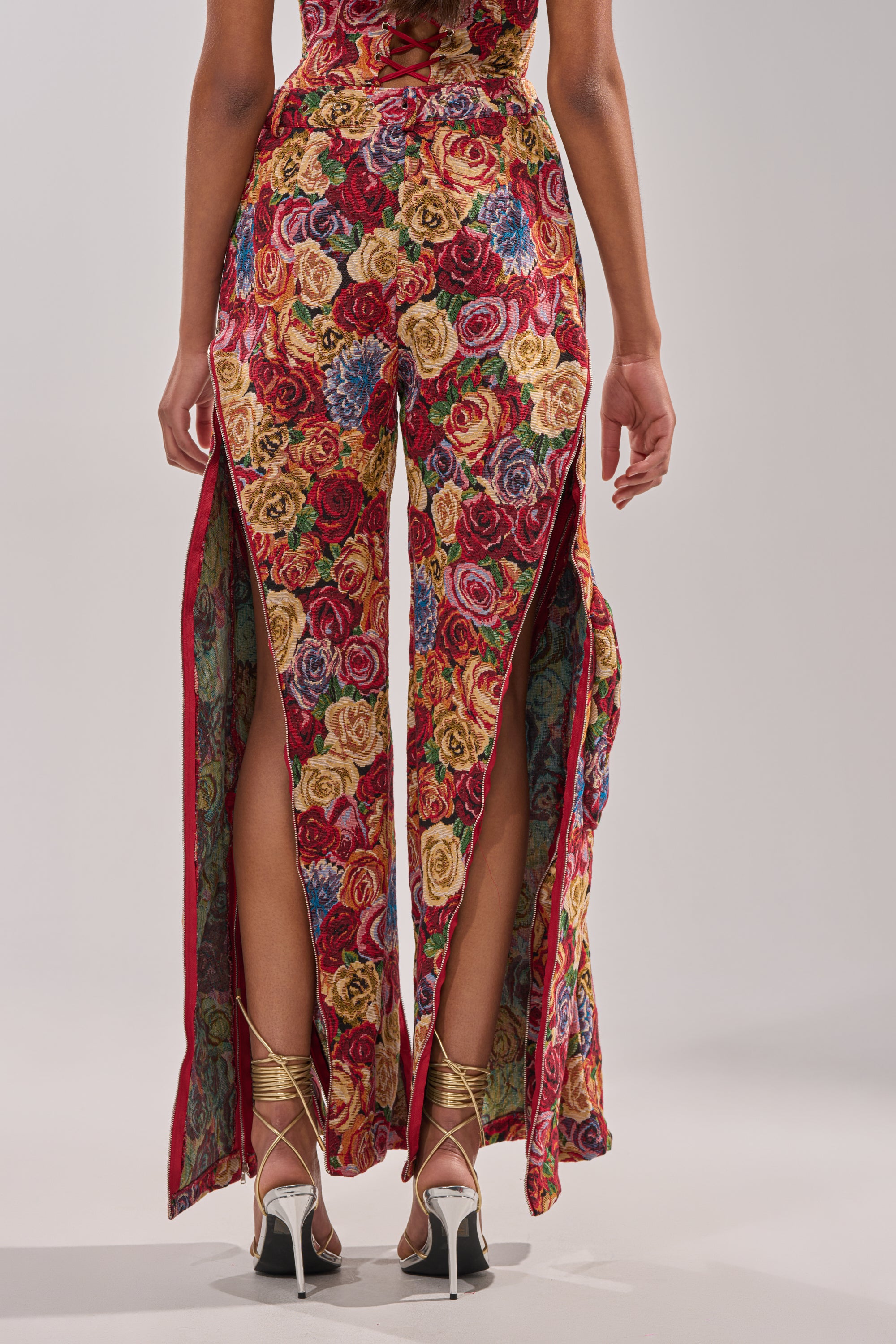 A person faces away wearing the INTO THE THICK OF IT FLORAL CARGO PANT, featuring vibrant floral tapestry, high side slits, and gold lace-up heels against a plain light background. 4