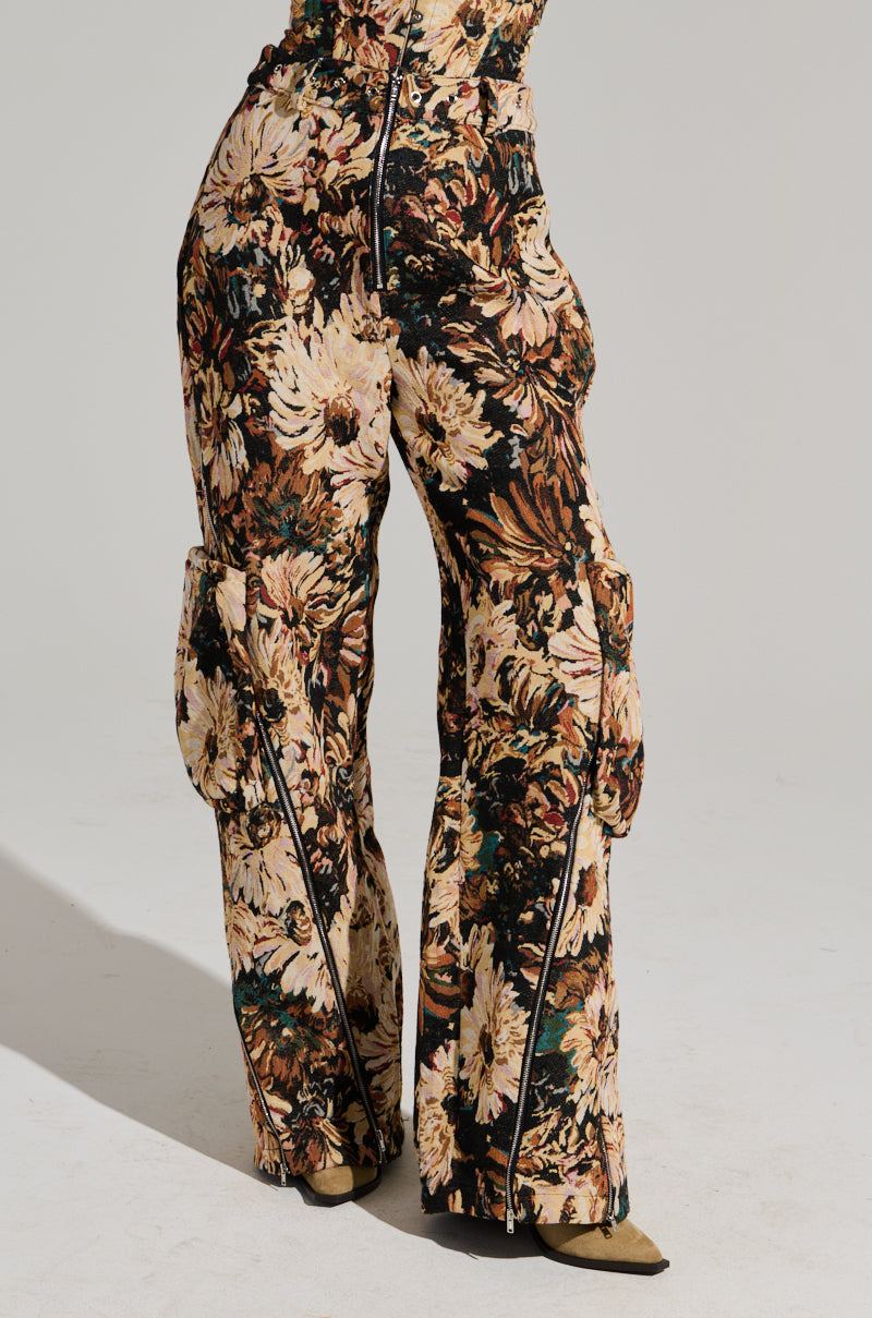 A person stands on a plain, light-colored background, donning the Fantasy Captured Cargo Pant in a vintage floral pattern with earthy tones. These high-waisted pants feature a wide-leg silhouette and come equipped with large side pockets and zippers running down the front of each leg.