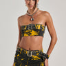 A person is wearing the TAKE IT FROM ME BANDEAU TOP in a yellow and black pattern, paired with a matching mini skirt adorned with silver eyelet details. They have a stylish hairstyle, hoop earrings, and a chunky necklace featuring a large spherical pendant, posing against a neutral background.