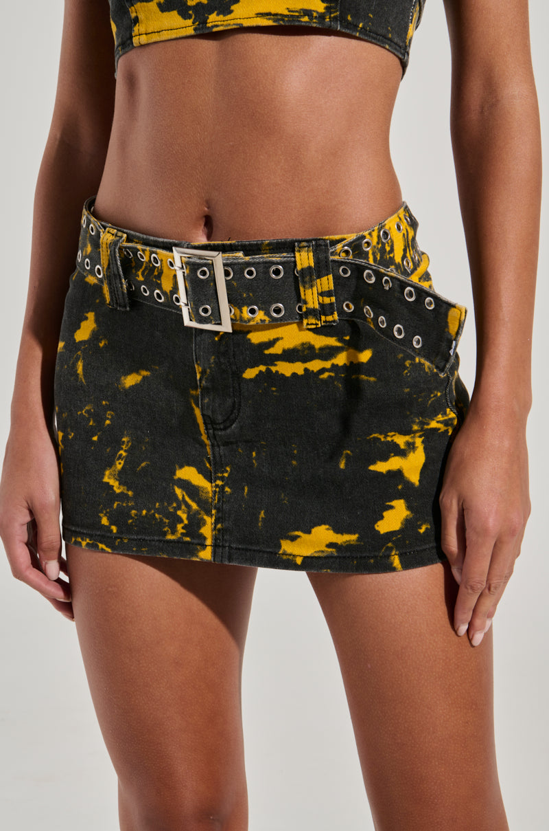 A person is wearing the "TAKE IT FROM ME MINI SKIRT," a black and yellow tie-dye mini skirt featuring a wide, adjustable belt with silver eyelets and a rectangular buckle. Their midriff is visible along with part of their top that matches the skirt.