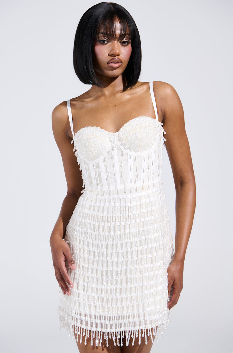 Against a plain background, someone in the SHOW STOPPER BEADED MINI DRESS with spaghetti straps and structural boning stands. The dress boasts intricate details and a textured design. They have straight hair and pose with one hand on their hip.