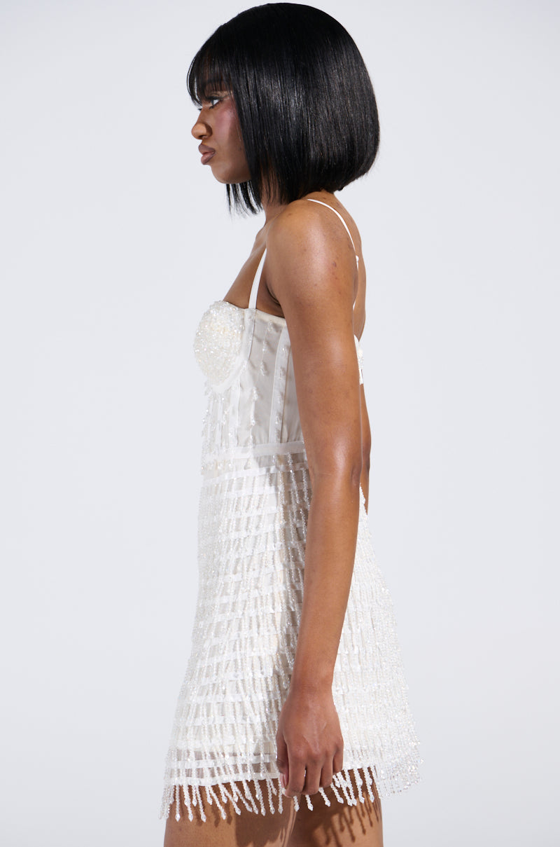 With shoulder-length black hair, a person wears the SHOW STOPPER BEADED MINI DRESS, a sleeveless white dress with a sweetheart neckline adorned in beads. They stand in profile against a plain background, highlighting the elegance of its beaded design.