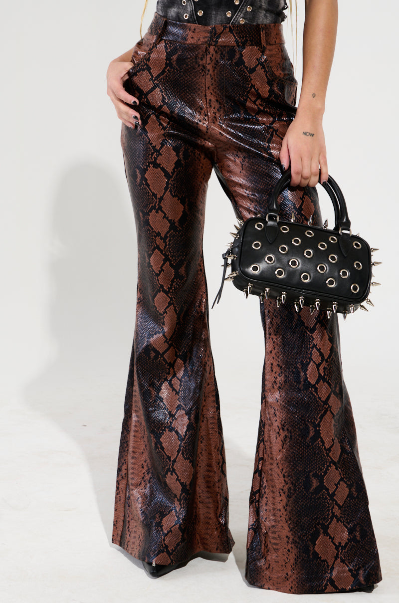 Clad in glimmering SNAKESKIN PRINCESS PU TROUSER, a person showcases high-rise style while holding a black handbag embellished with metal spikes and grommets, achieving an impeccable faux leather chic. The backdrop is simple and light-colored.