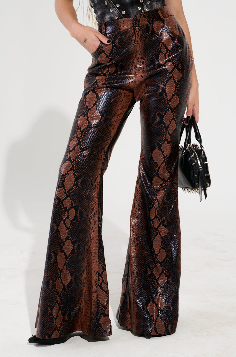 A person wearing SNAKESKIN PRINCESS PU TROUSER with a stylish high-rise design and a black faux leather top holds a studded black handbag.