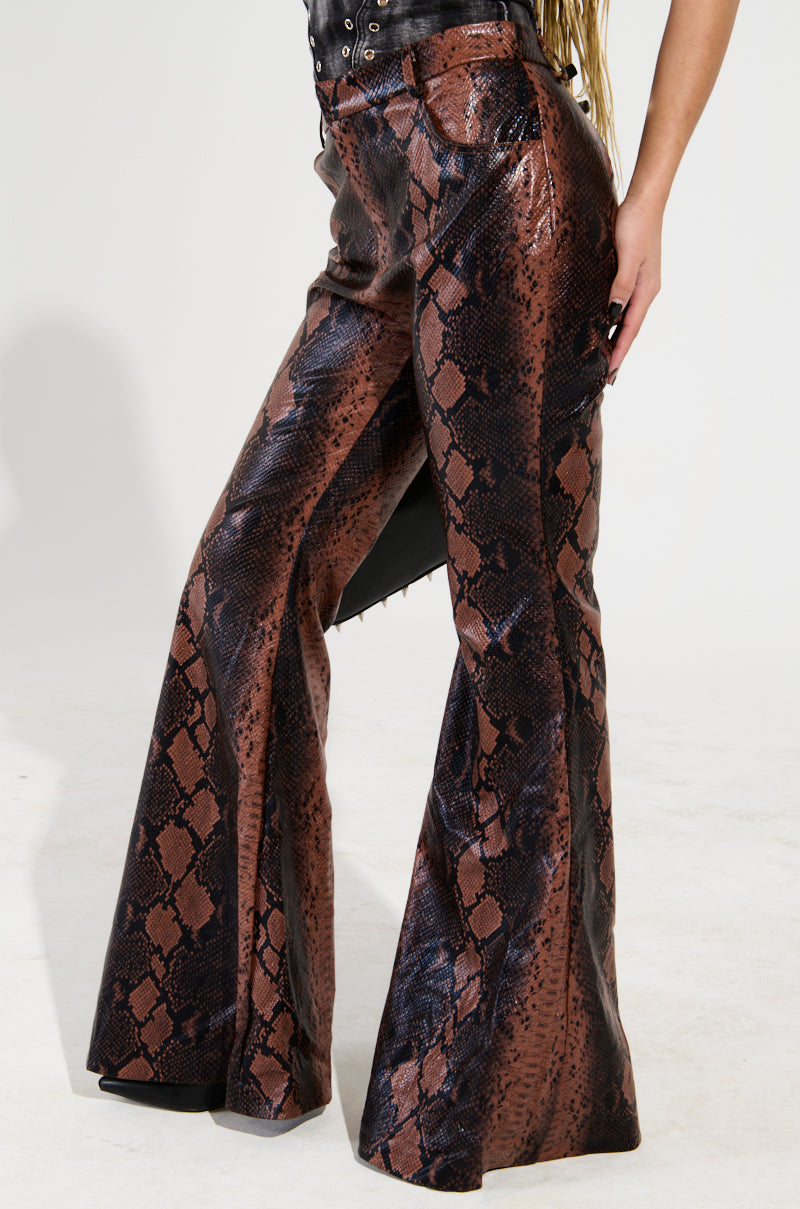 A person wearing the SNAKESKIN PRINCESS PU TROUSER, featuring a high-waisted, flared design with a brown and black snakeskin pattern. The trousers are crafted from shiny, textured faux leather. A hint of a black top is visible, set against a neutral background.