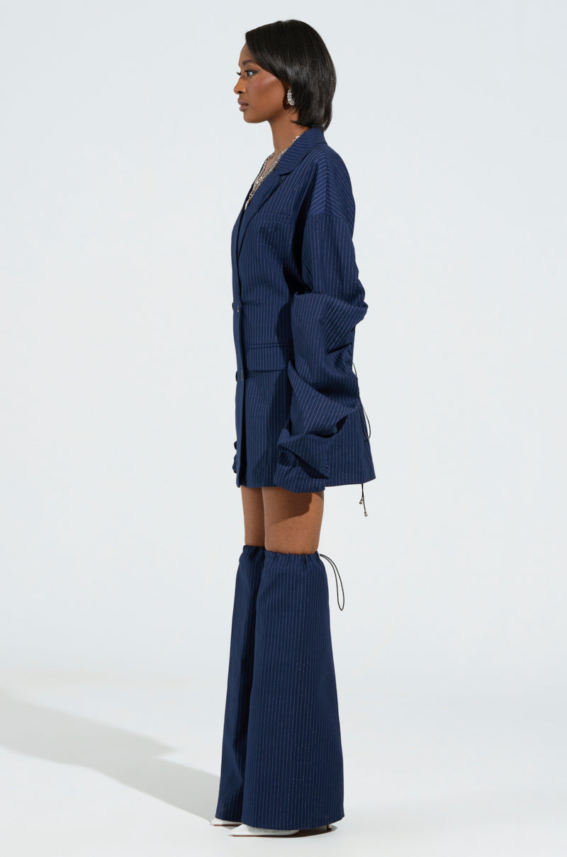 Dressed in the BUSINESS MEETING CHIC BLAZER DRESS AND LEG WARMER SET, a person poses against a plain backdrop. The ensemble, highlighted by exaggerated sleeves and distinctive pant design, creates a stylish and bold silhouette with its oversized, elegant navy blue pinstripe blazer paired with matching wide-leg pants and a faux leather corset. 2