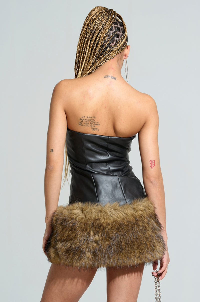 A person with braided hair is facing away, showcasing the BARON PU MINI DRESS WITH FUR TRIM, a strapless sleek faux leather dress featuring a fur trim on the skirt. They have visible tattoos on their back and arm against a plain, light-colored background.