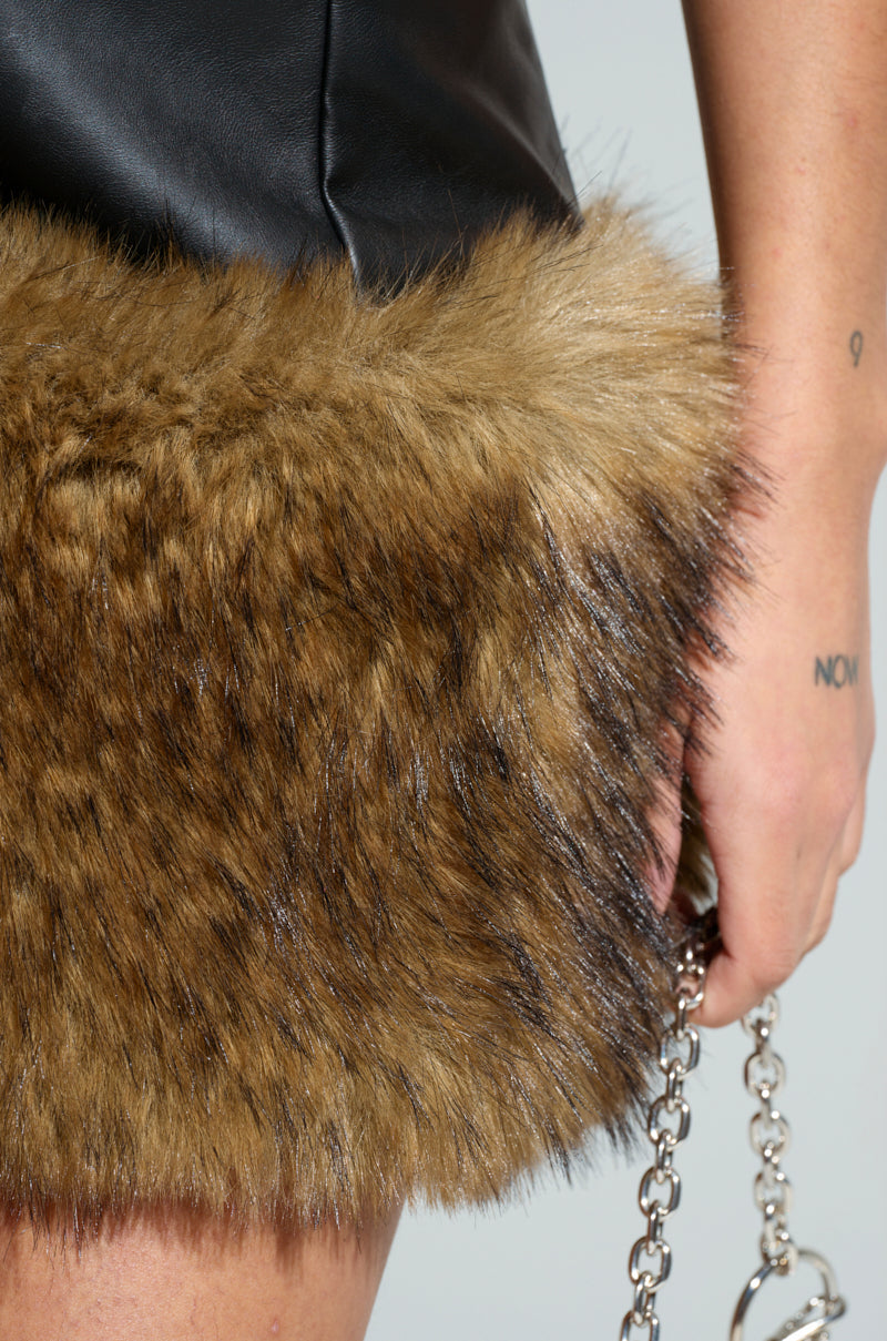 A close-up of a person wearing the BARON PU MINI DRESS WITH FUR TRIM reveals the intricate details of the black faux leather and brown fur trim. Their tattooed hand grips a silver chain, accentuating the texture of the materials.