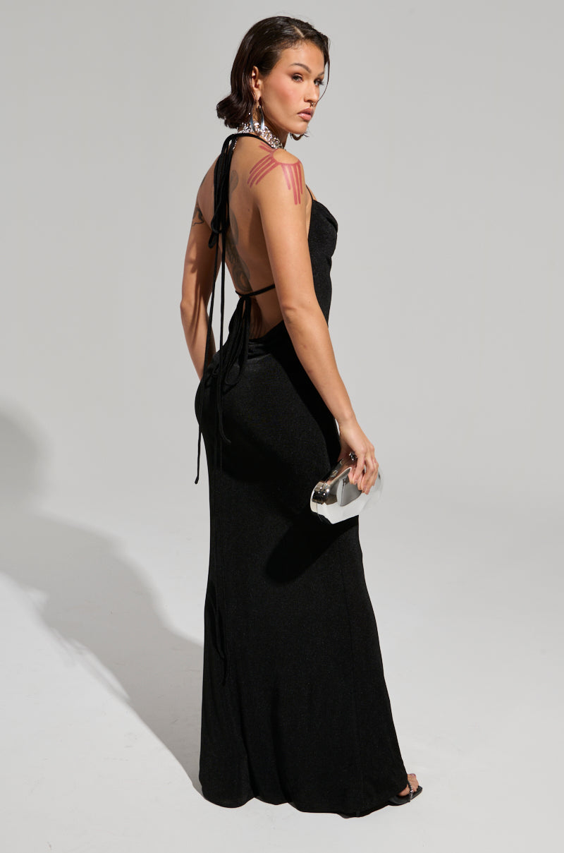 A person in the FOR EVERY OCCASION SLINKY MAXI DRESS with an open back design stands facing away, holding a silver clutch. Their short hair reveals visible tattoos on their arm against the plain background.