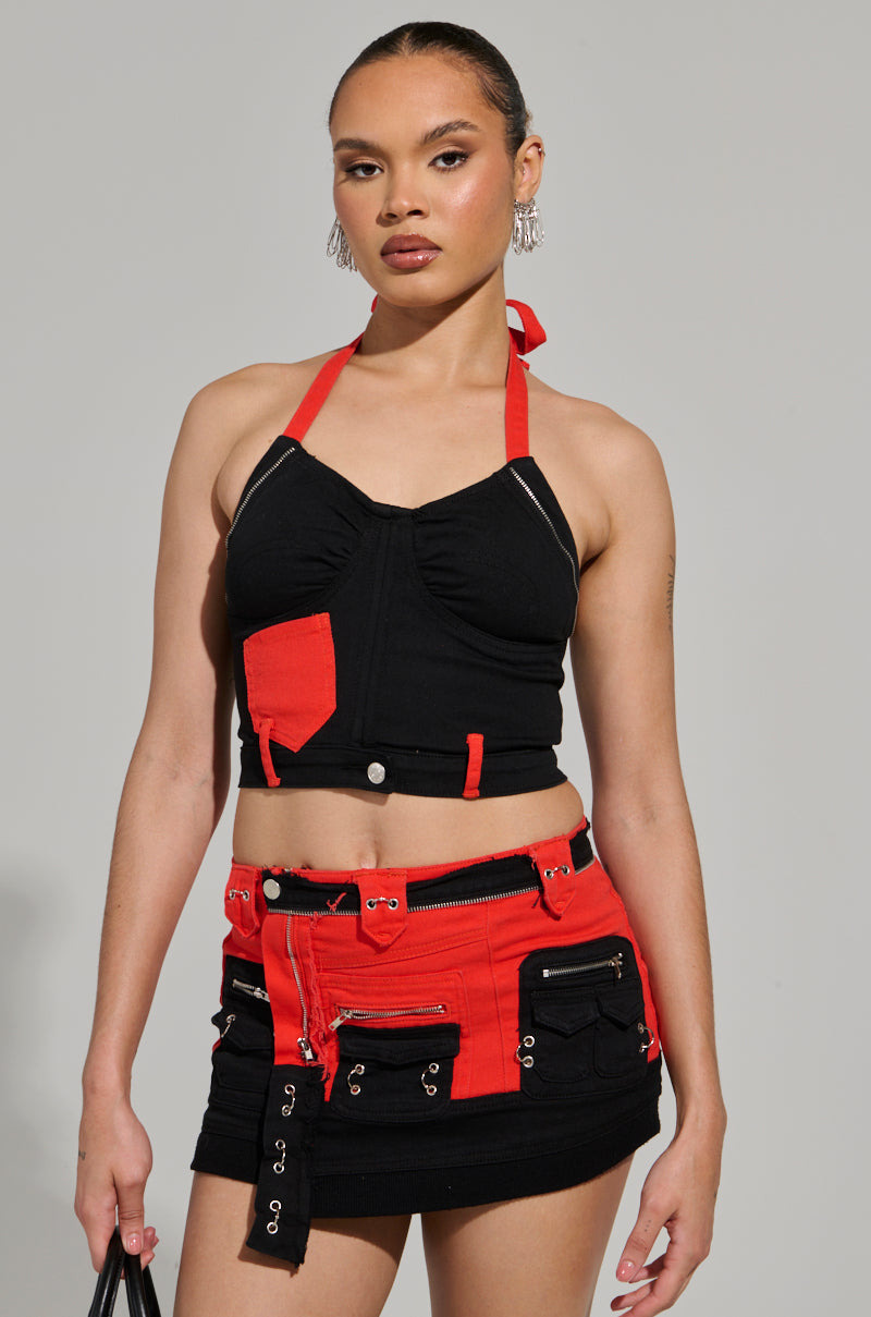 A person stands confidently in the Motor Sport Corset Top in Black & Red, paired with a matching mini skirt featuring zippers and pockets. They hold a black bag, exuding a neutral expression against a plain background.